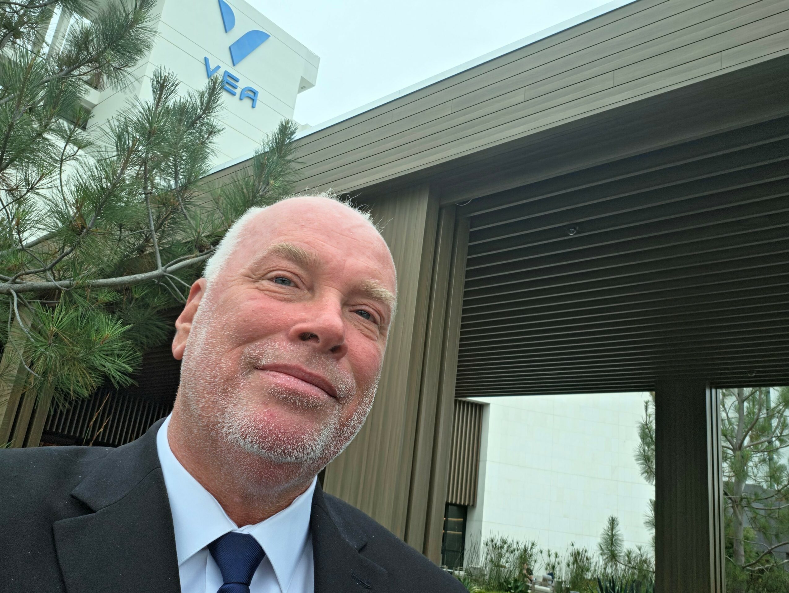 Man with white hair and beard smiling outdoors near a building with trees in the background.