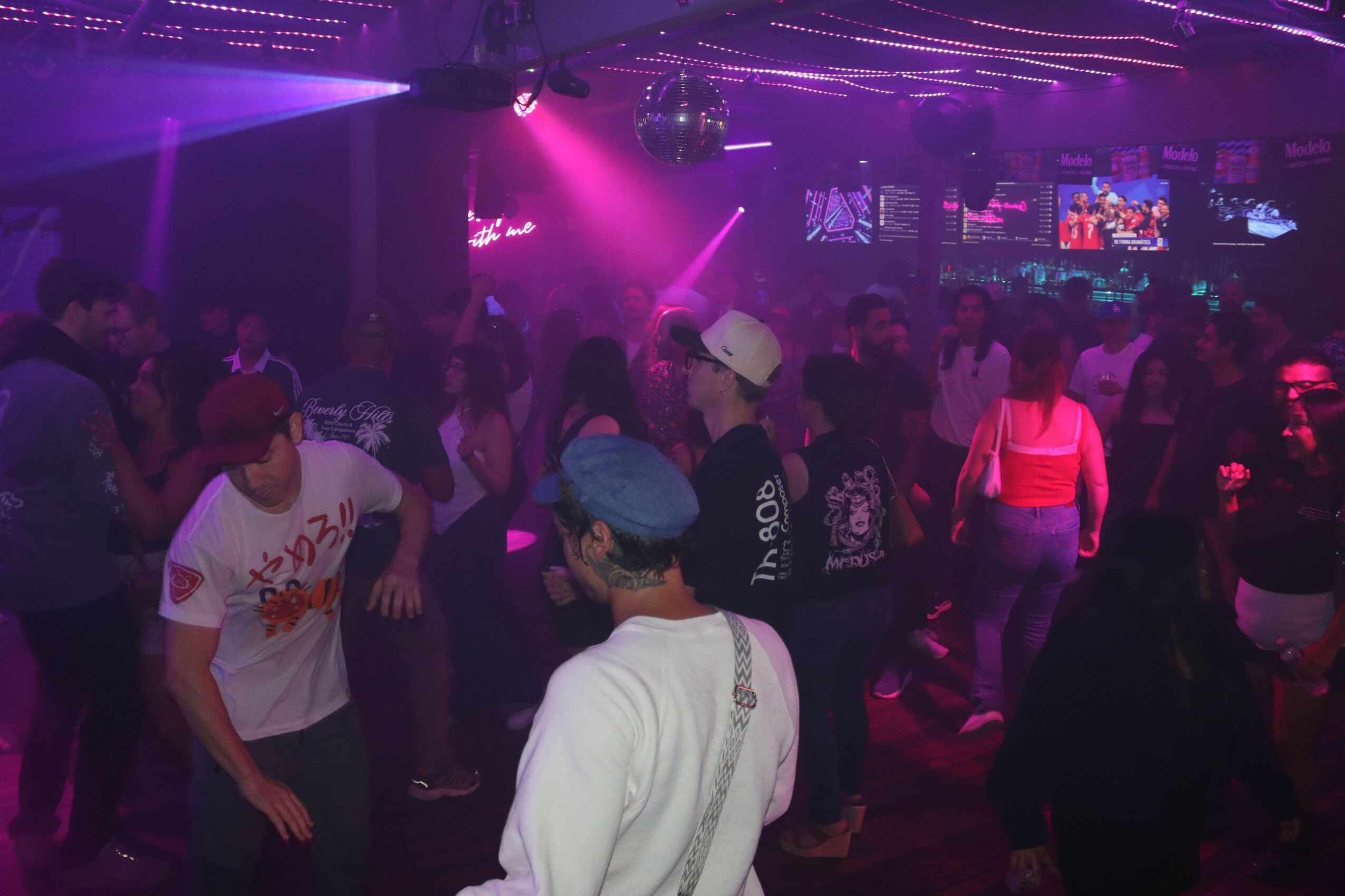 People dancing in a nightclub with purple and pink lighting, multiple screens, and a disco ball overhead.