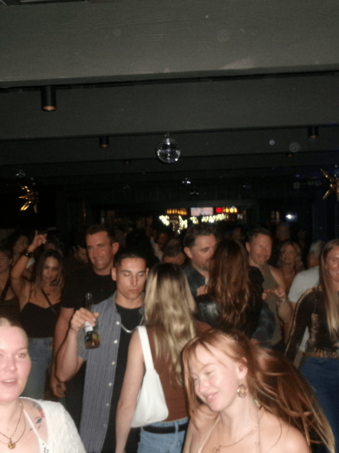 Crowd of people at a social event, some holding drinks, in a dimly lit indoor setting.