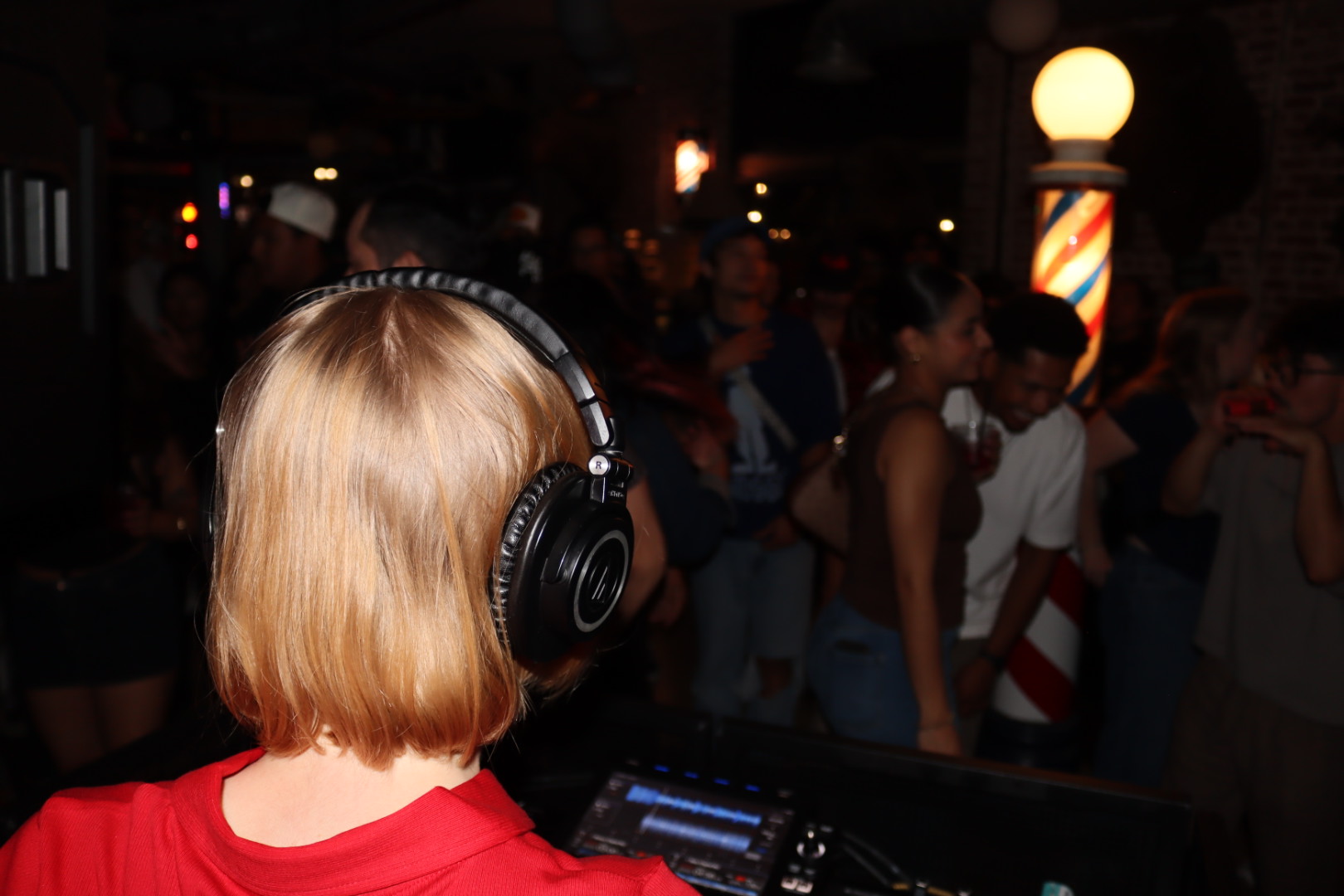 Person with blonde hair wearing headphones, DJ equipment, crowd in background at night, illuminated lamp post, lively atmosphere.