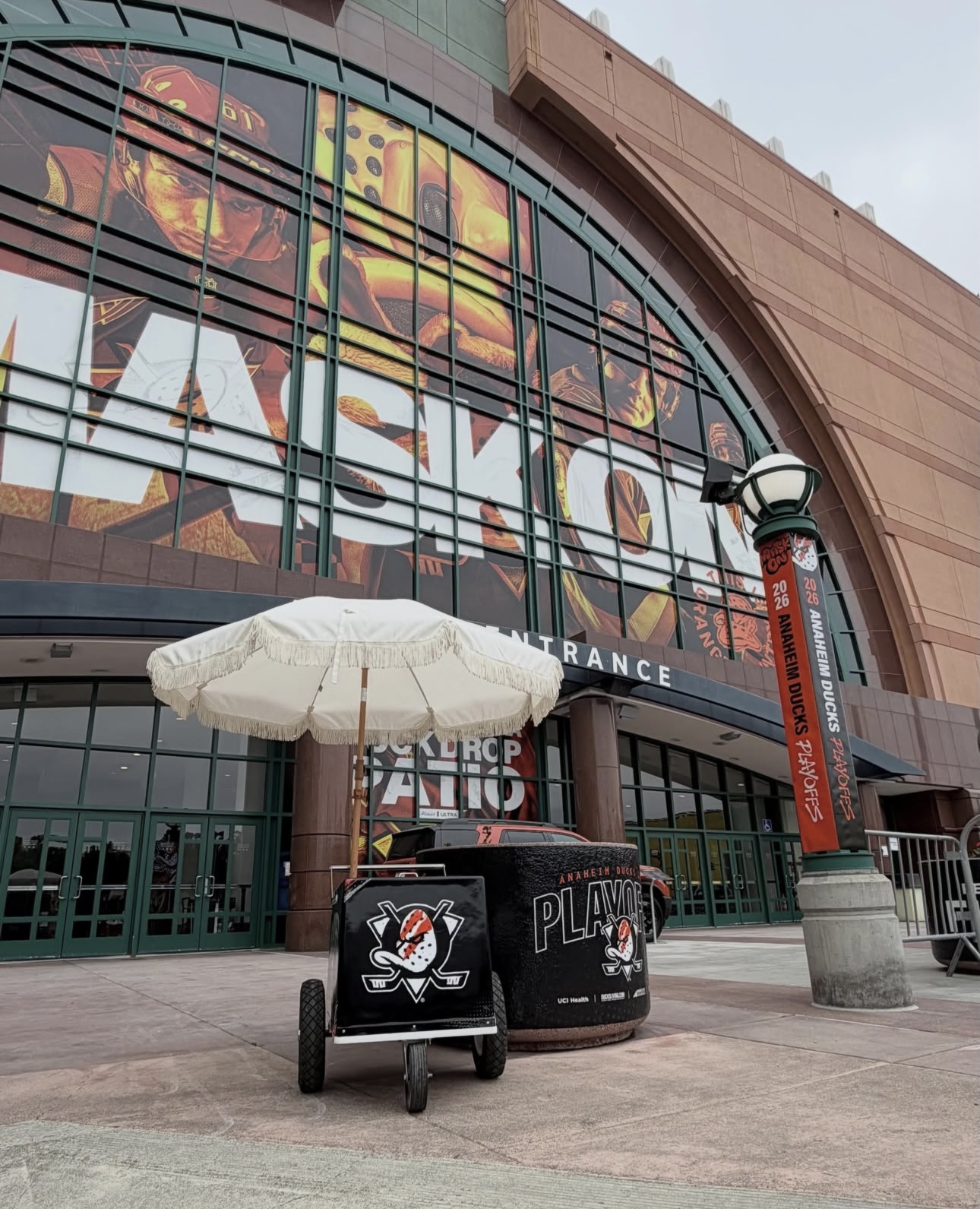 Entrance of a building with large glass windows and a sign reading 'PLAYOFFS'. A white umbrella, a black cart, and a trash bin are in front.
