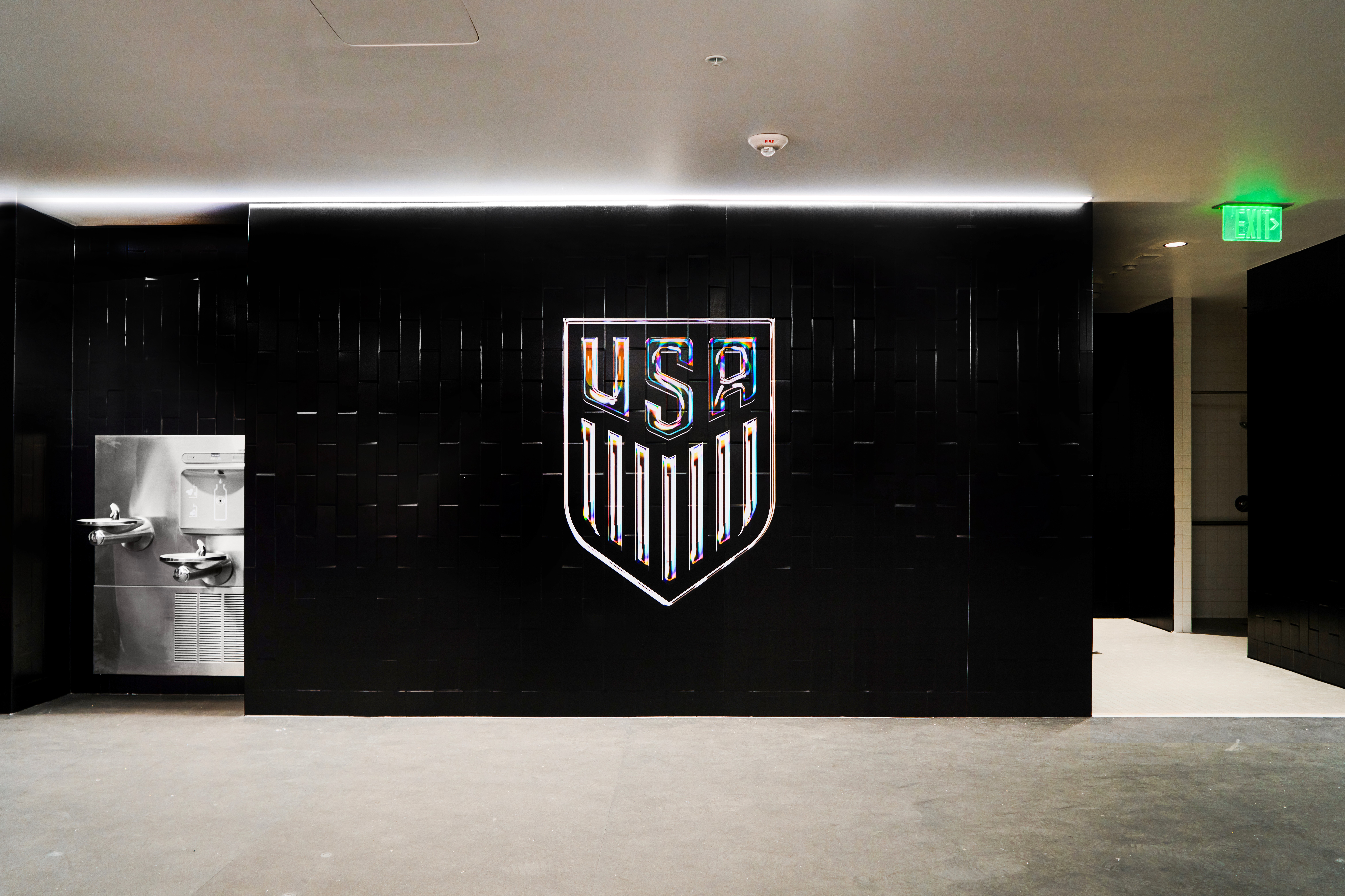 Black wall with a USA shield logo in the center, illuminated by overhead lighting, with a door on the right and a water fountain on the left.