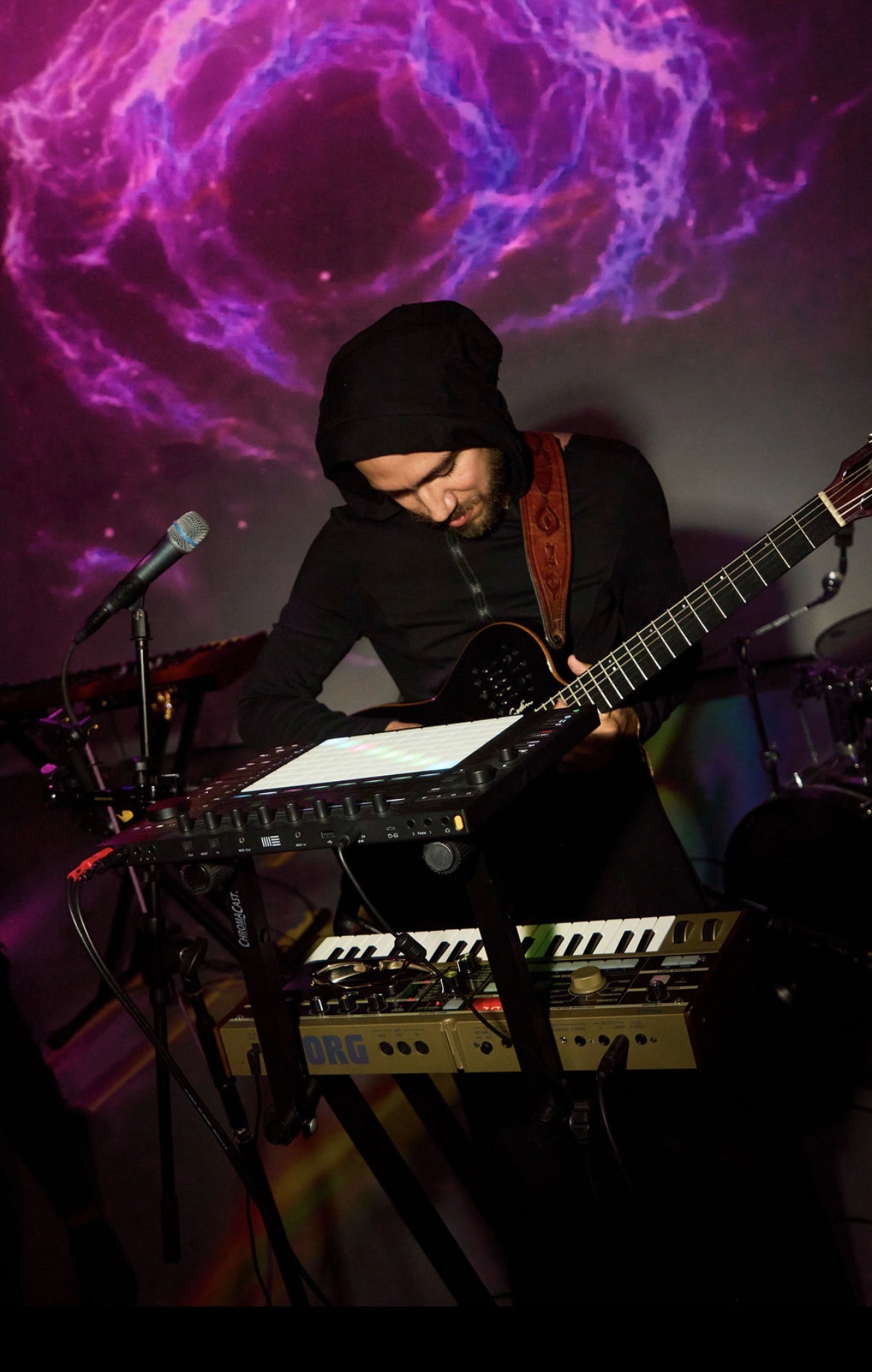 Musician wearing a black hoodie playing guitar and keyboard with a cosmic purple and pink background.