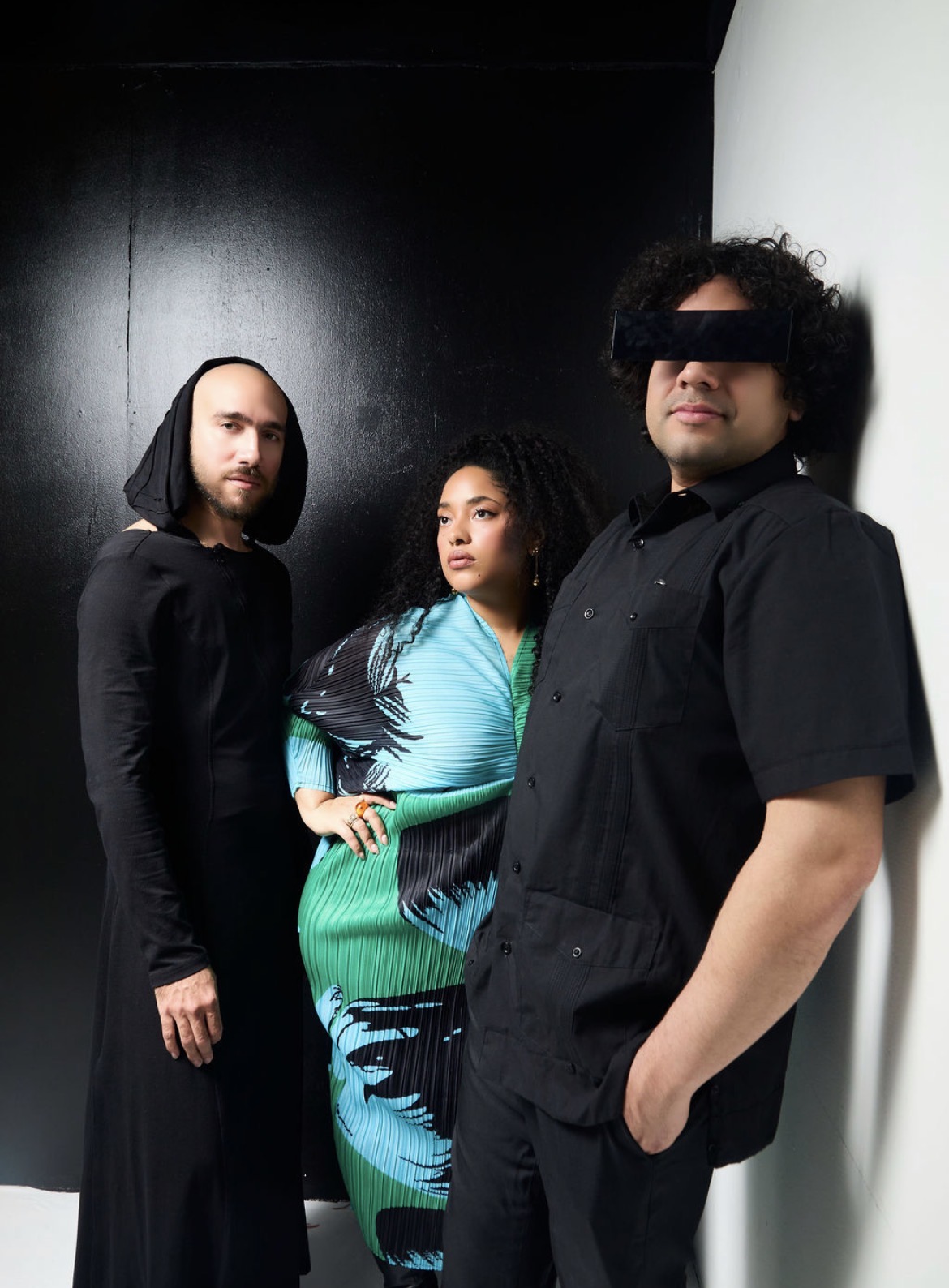 Three people standing indoors against a dark and white wall, posing for a photo, with one person wearing a black hooded cloak.