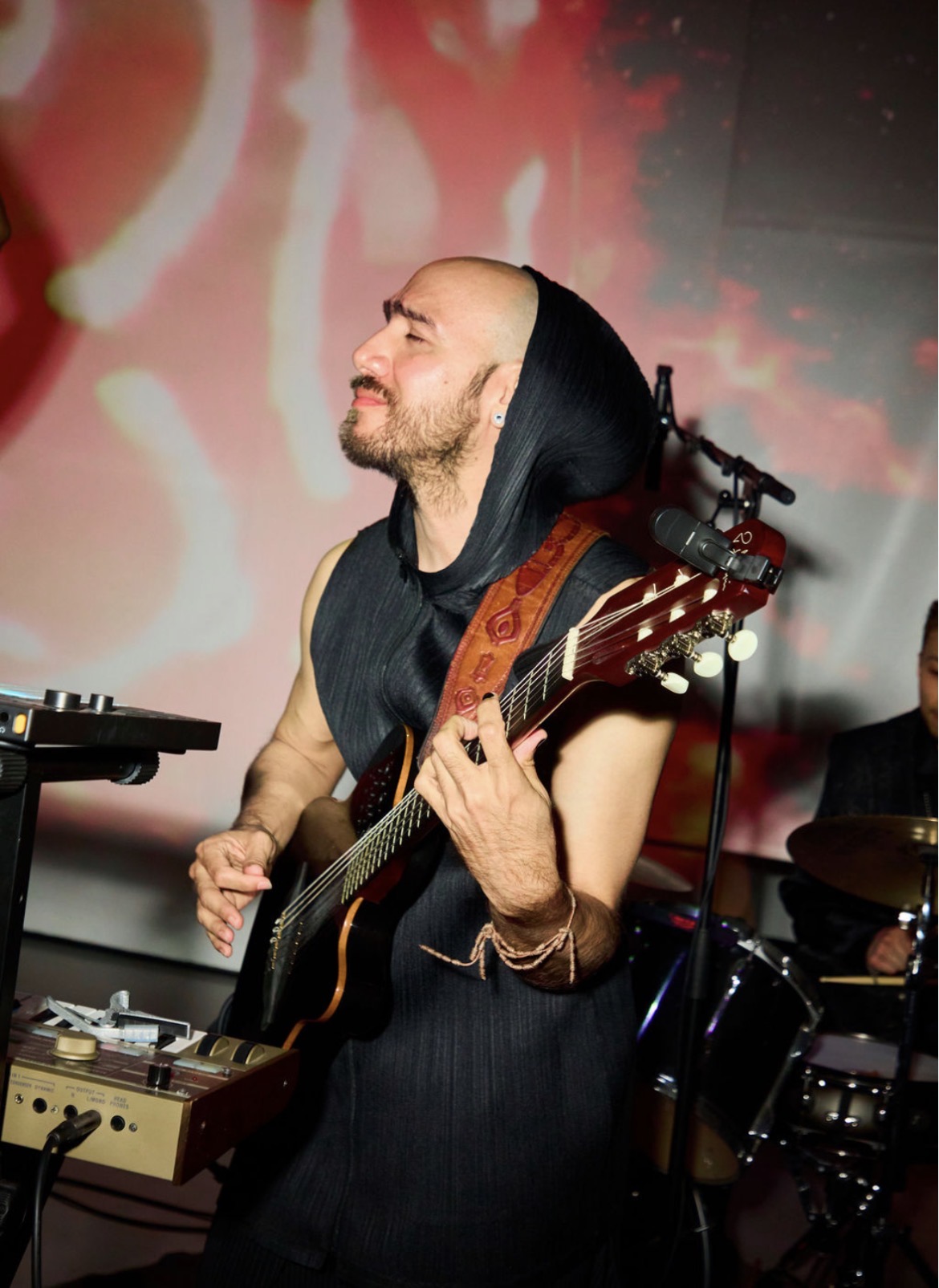 Person playing guitar with eyes closed, wearing a hooded garment, on stage with colorful background.