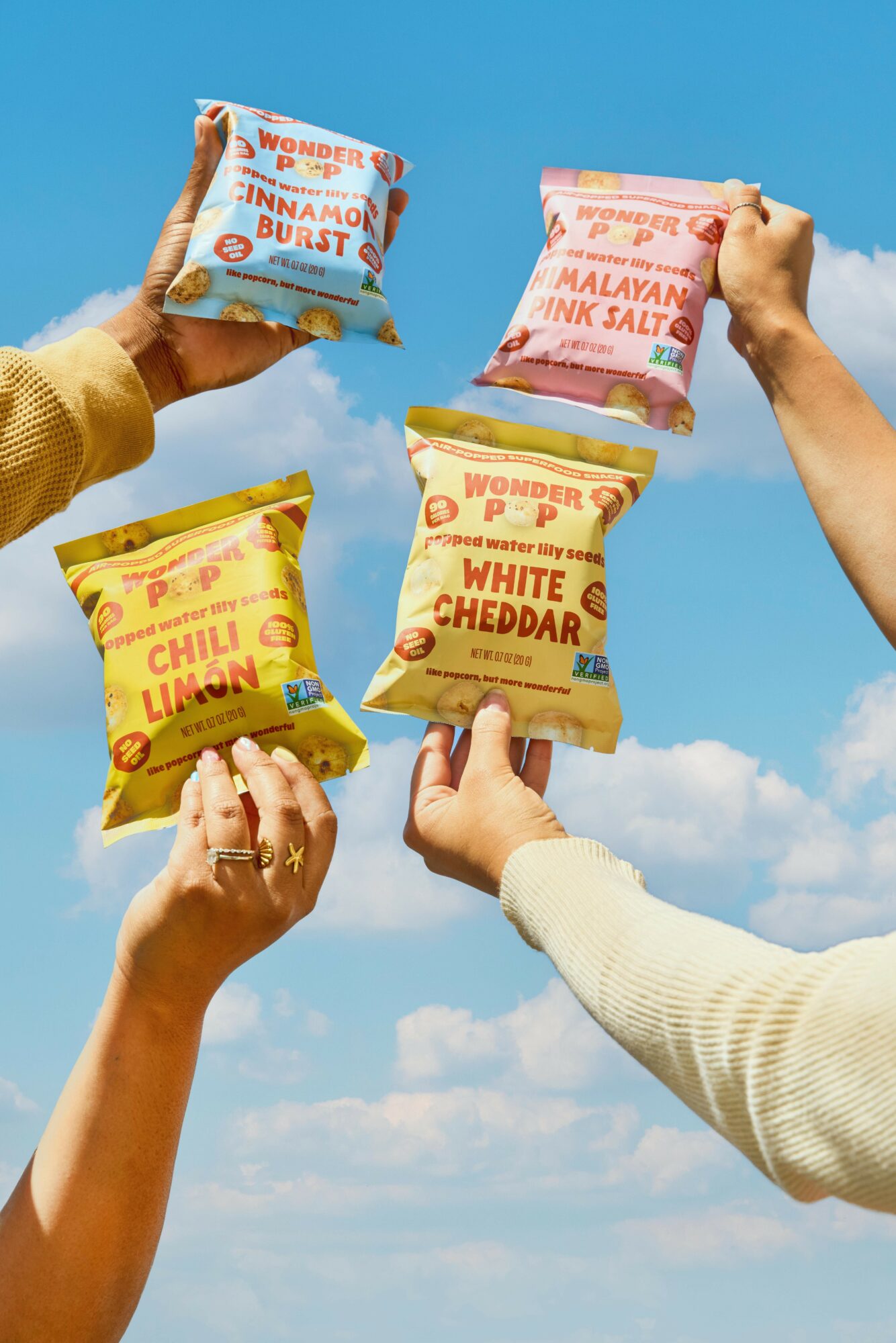 Four hands hold different snack bags against a blue sky with clouds, each bag has colorful text and graphics.