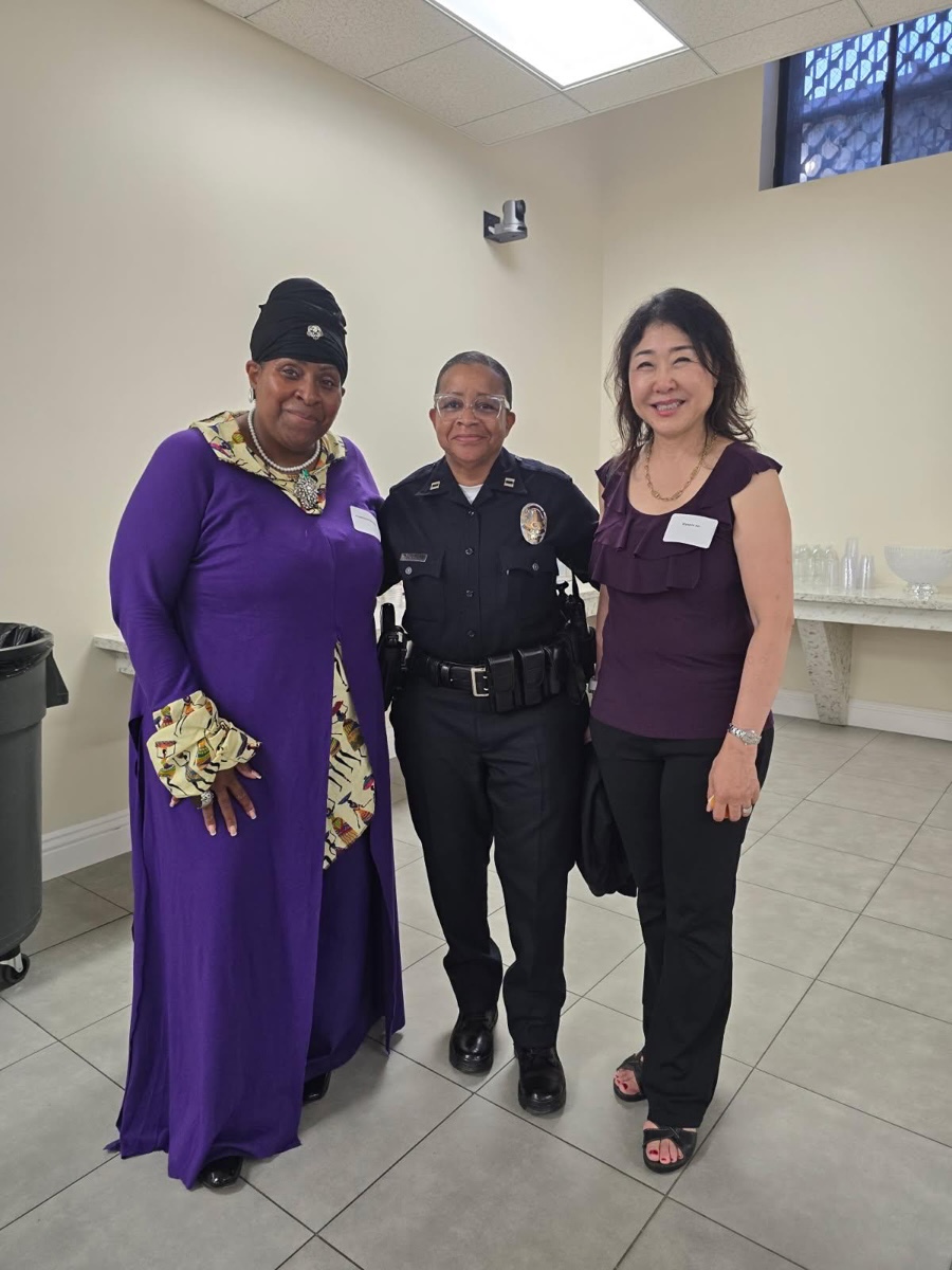 Three women stand together indoors, one in a purple dress, one in a police uniform, and one in a purple top and black pants.