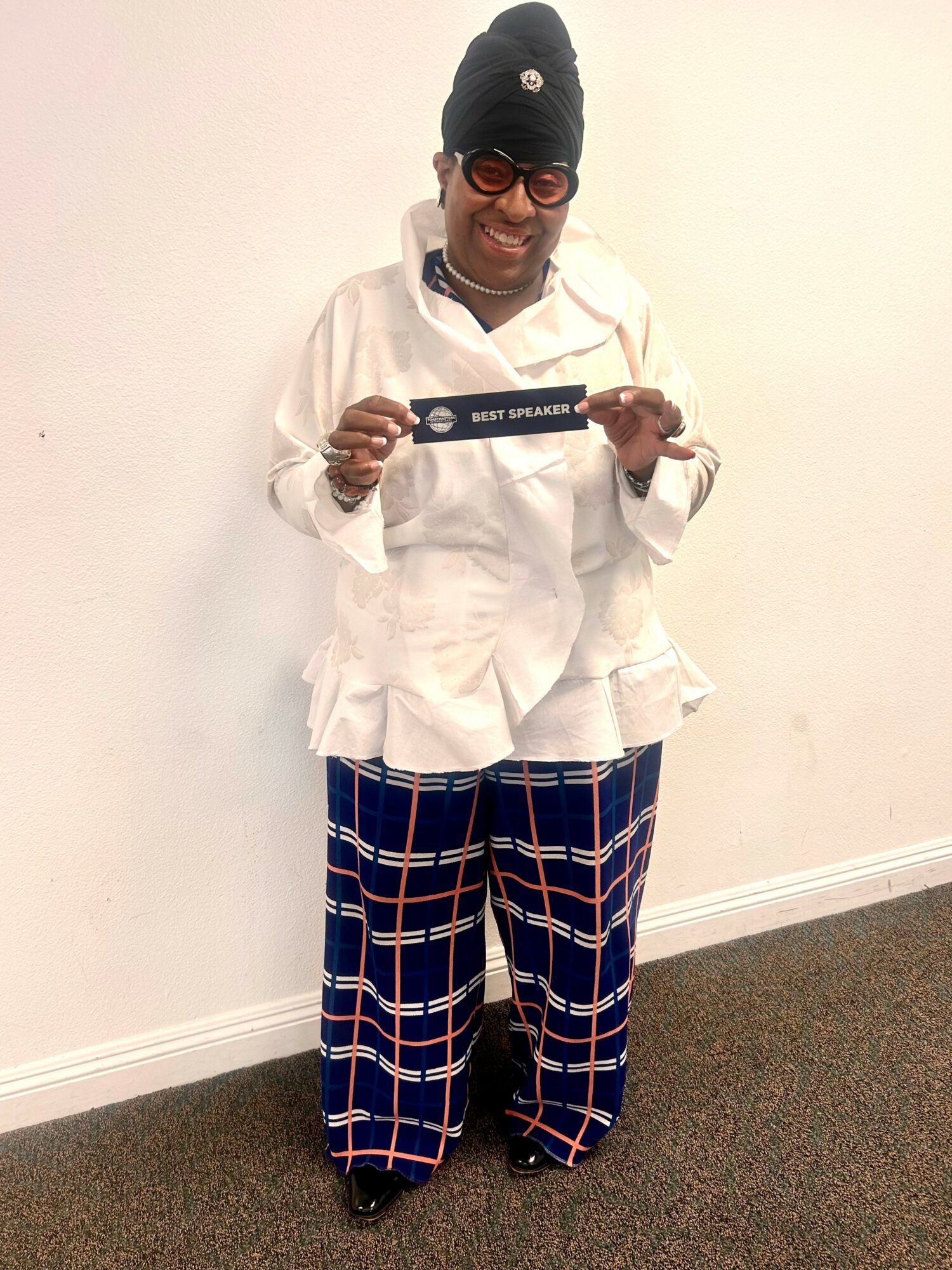 Person smiling, wearing glasses, a beanie, white coat, and plaid pants, holding a sign, standing against a plain wall.