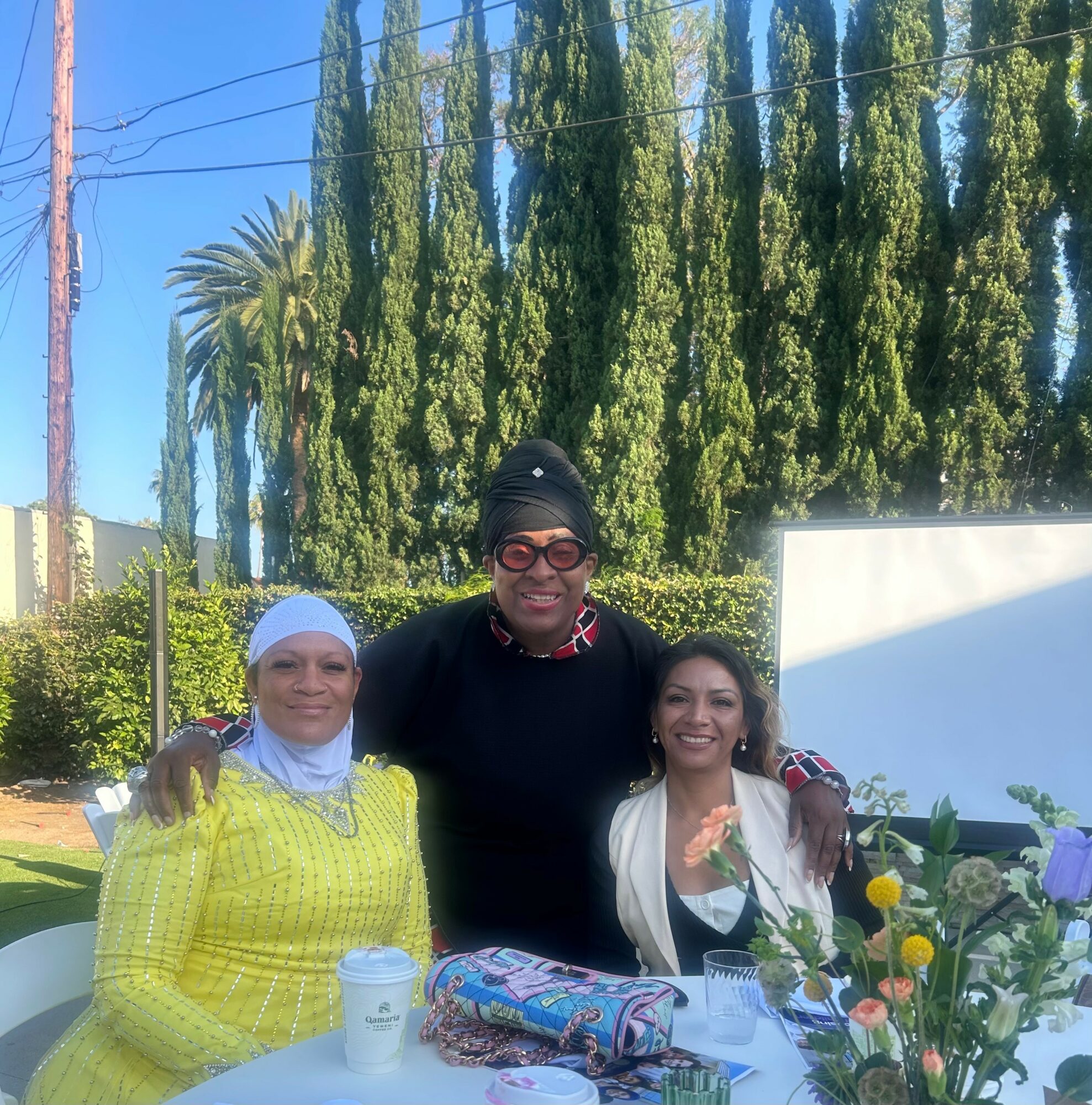 Three women standing outdoors with trees and a white wall in the background, smiling, with flowers and a bag in front.