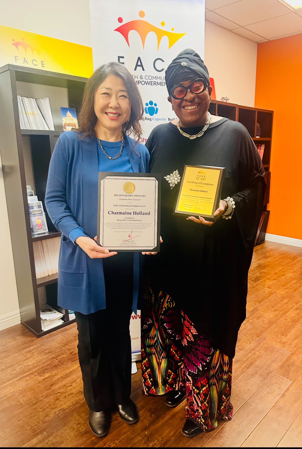 Two women standing indoors, smiling, holding awards, with a banner and bookshelf in the background.