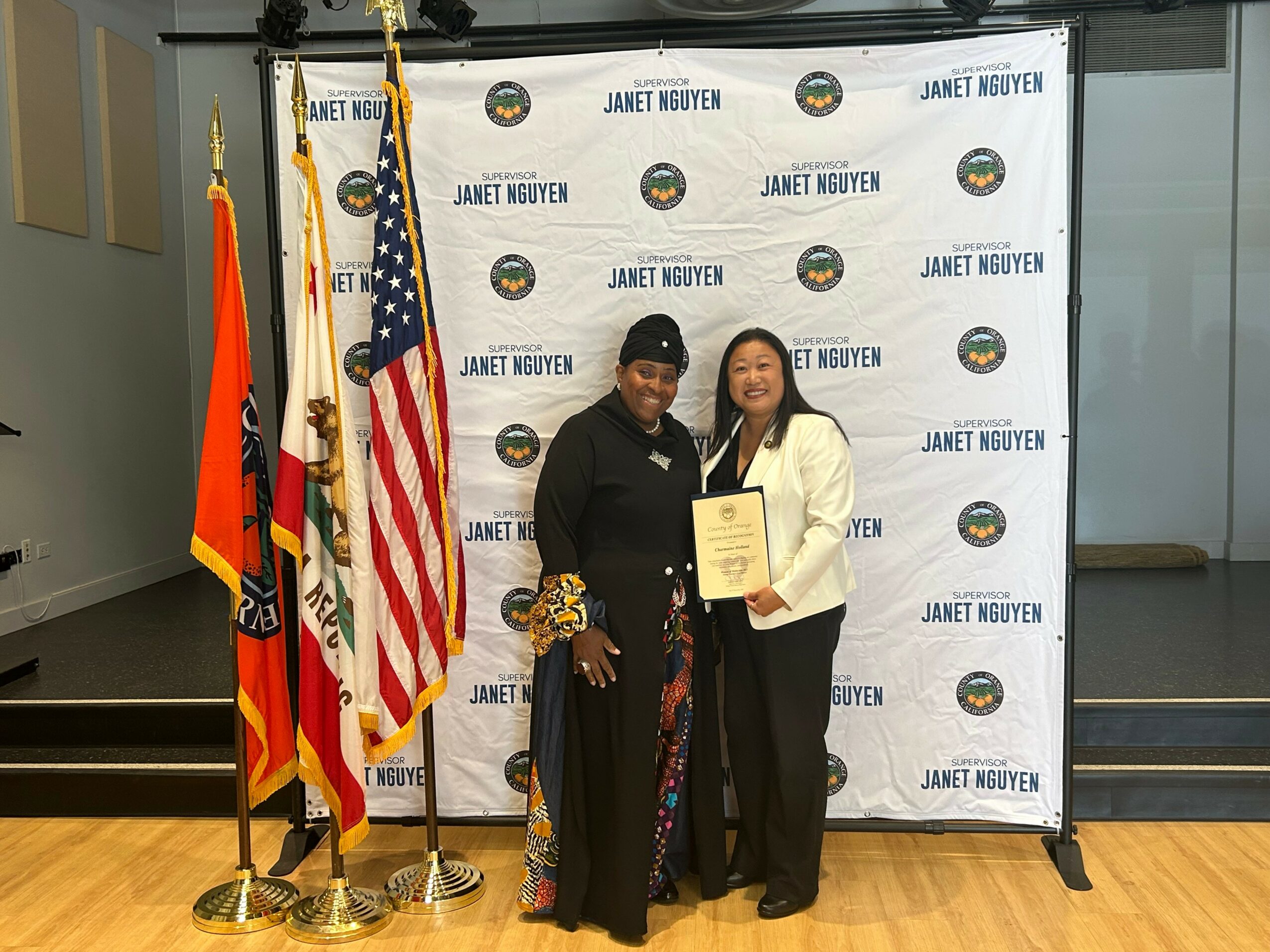 Two women stand in front of a backdrop with flags, holding a certificate, smiling. One woman wears a black outfit, the other a white blazer.