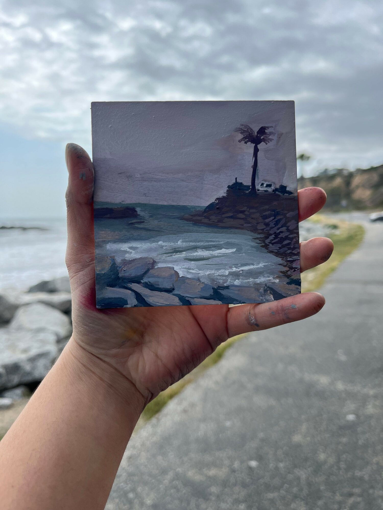 Person holding a small painting of a seascape with rocks and waves, outdoors near water and rocks.