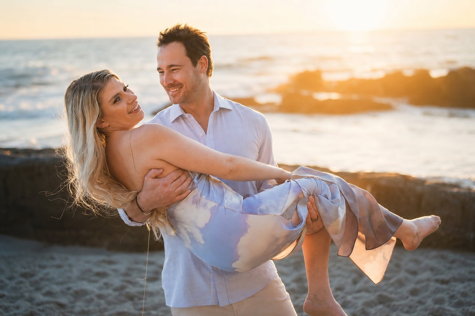 A couple at the beach during sunset, the man holding the woman in his arms, both smiling.