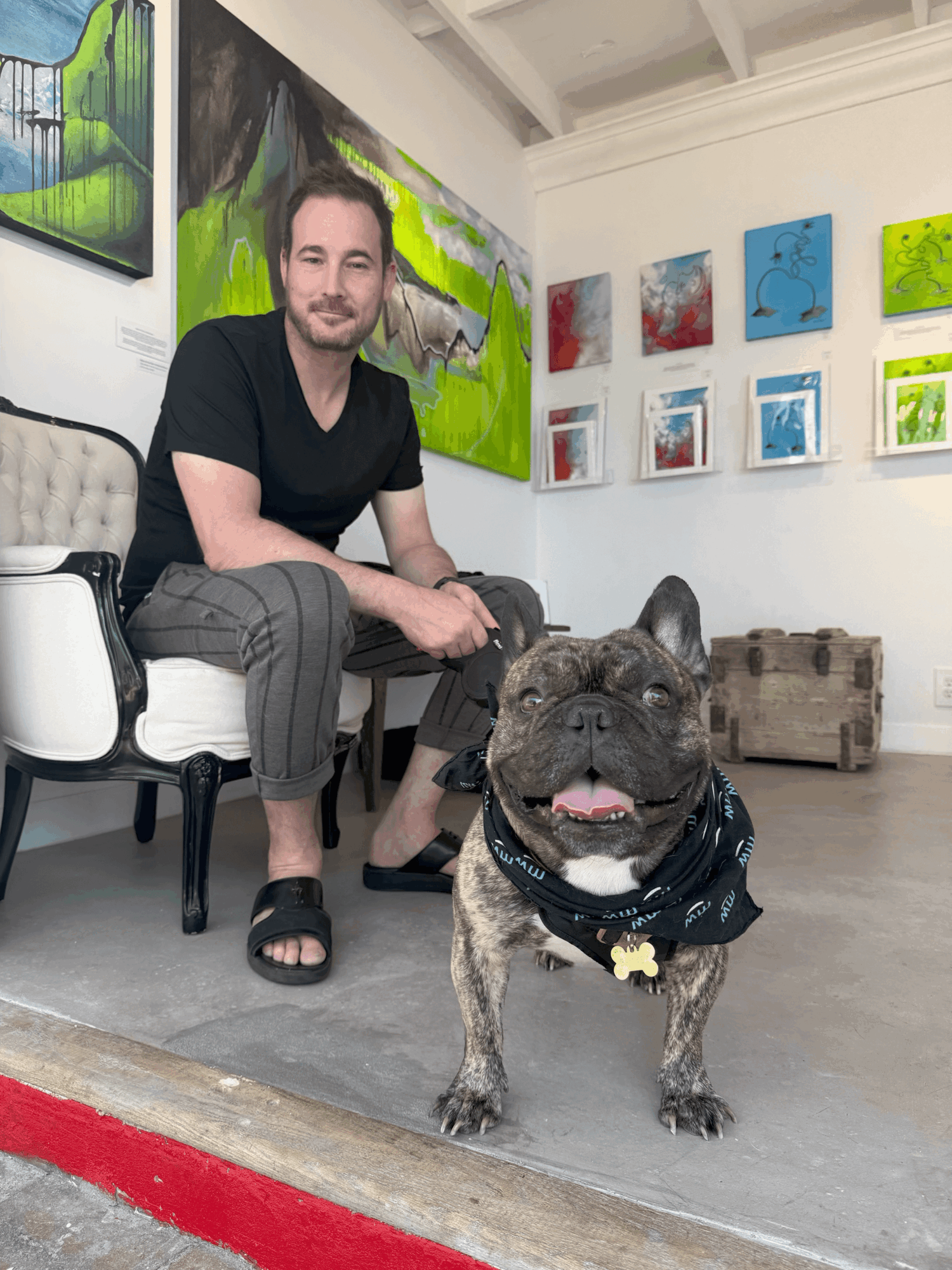 Man sitting on a chair with a French Bulldog in an art gallery, colorful paintings on the wall behind them.