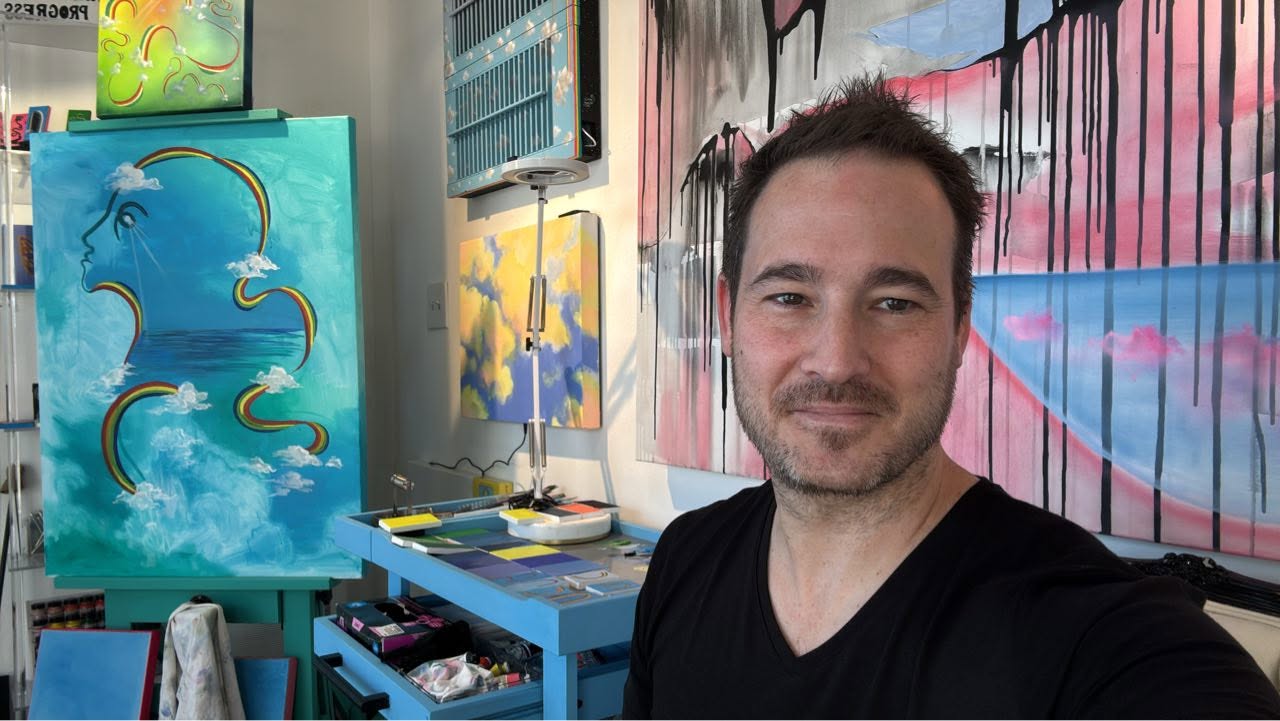 Man taking a selfie in an art studio with colorful paintings and supplies in the background.