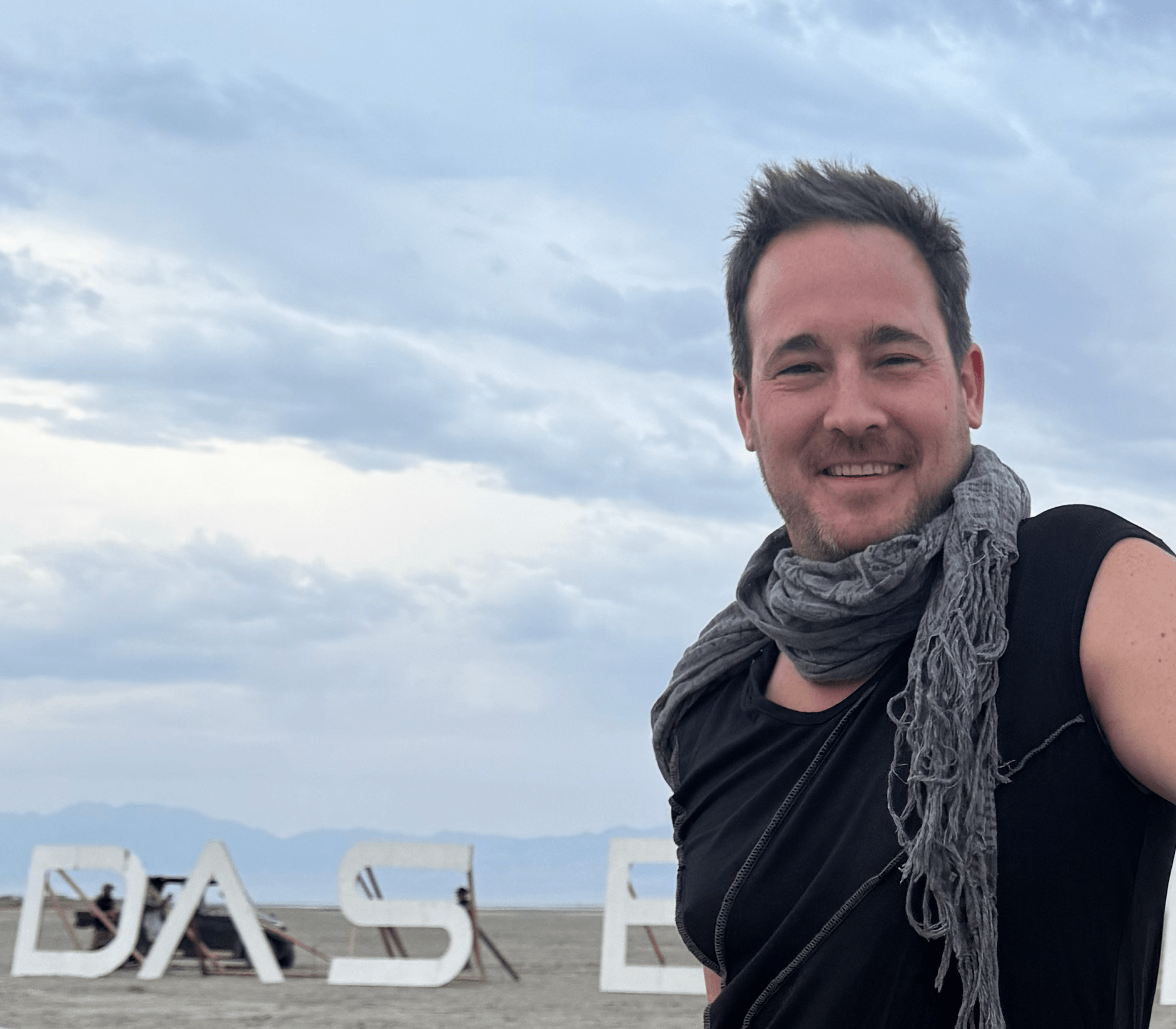 Smiling man with short dark hair and a gray scarf outdoors with cloudy sky and large white letters in background.