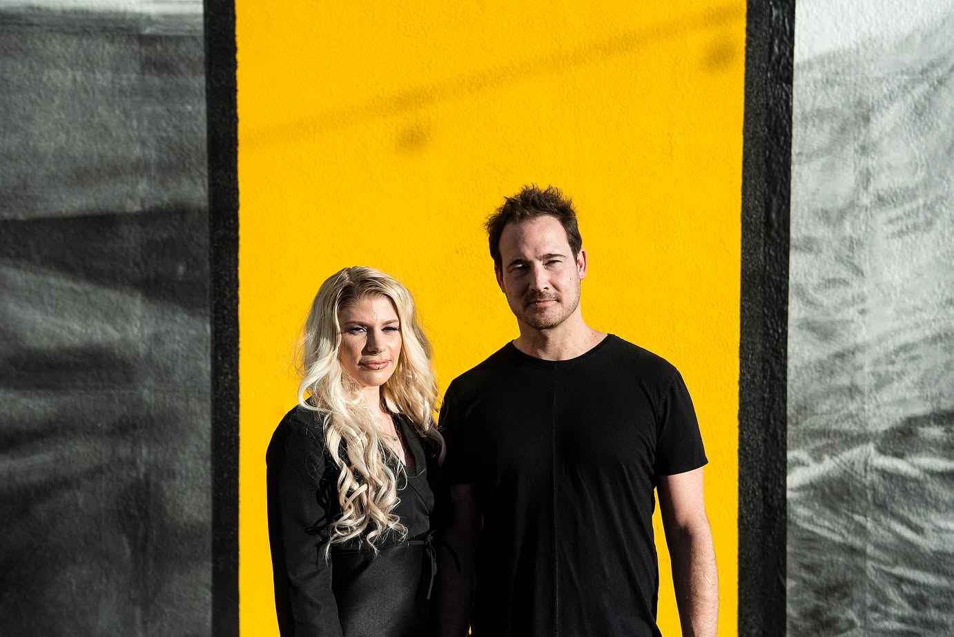 A man and woman stand side by side in front of a yellow and black background, smiling at the camera.