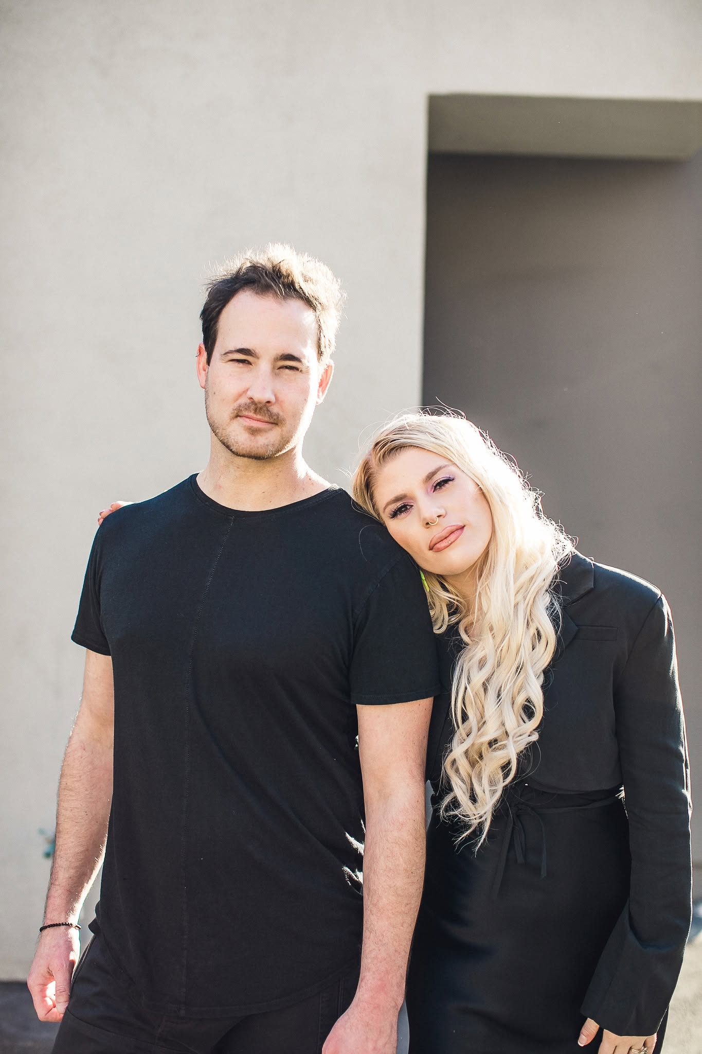A man and woman stand outdoors, smiling, with a plain wall and doorway in the background.