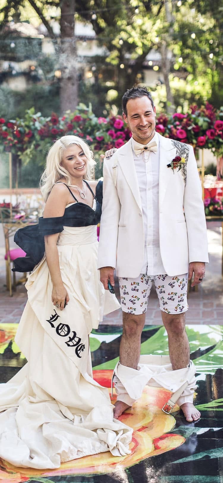 A woman in a black and white dress and a man in a white suit with shorts stand outdoors with colorful flowers and trees.