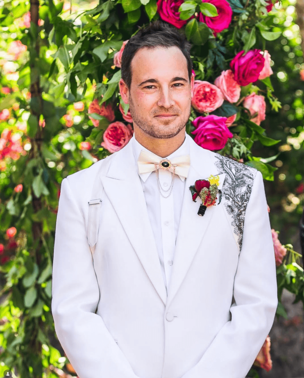 Man in white suit with boutonniere, standing in front of pink and red flowers, smiling.