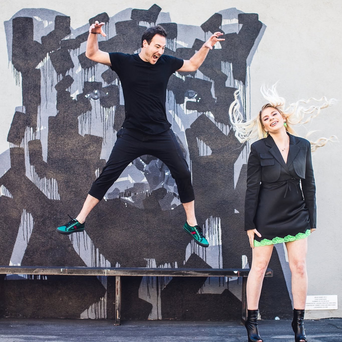 A man jumps with arms raised, and a woman stands smiling beside him against a graffiti wall.