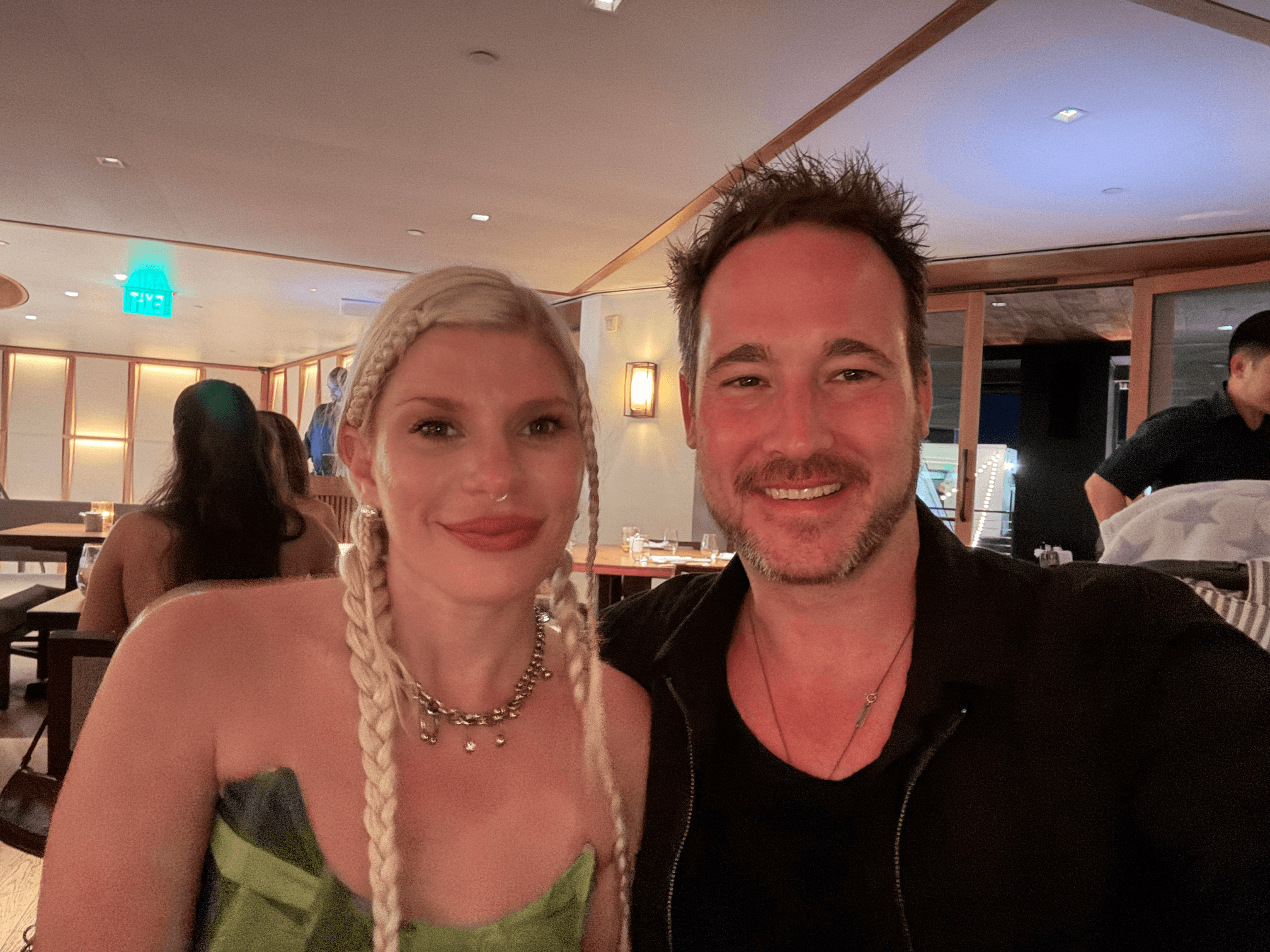 Two people smiling at a social event, woman with long braided blonde hair and man with short dark hair, in a warmly lit room.