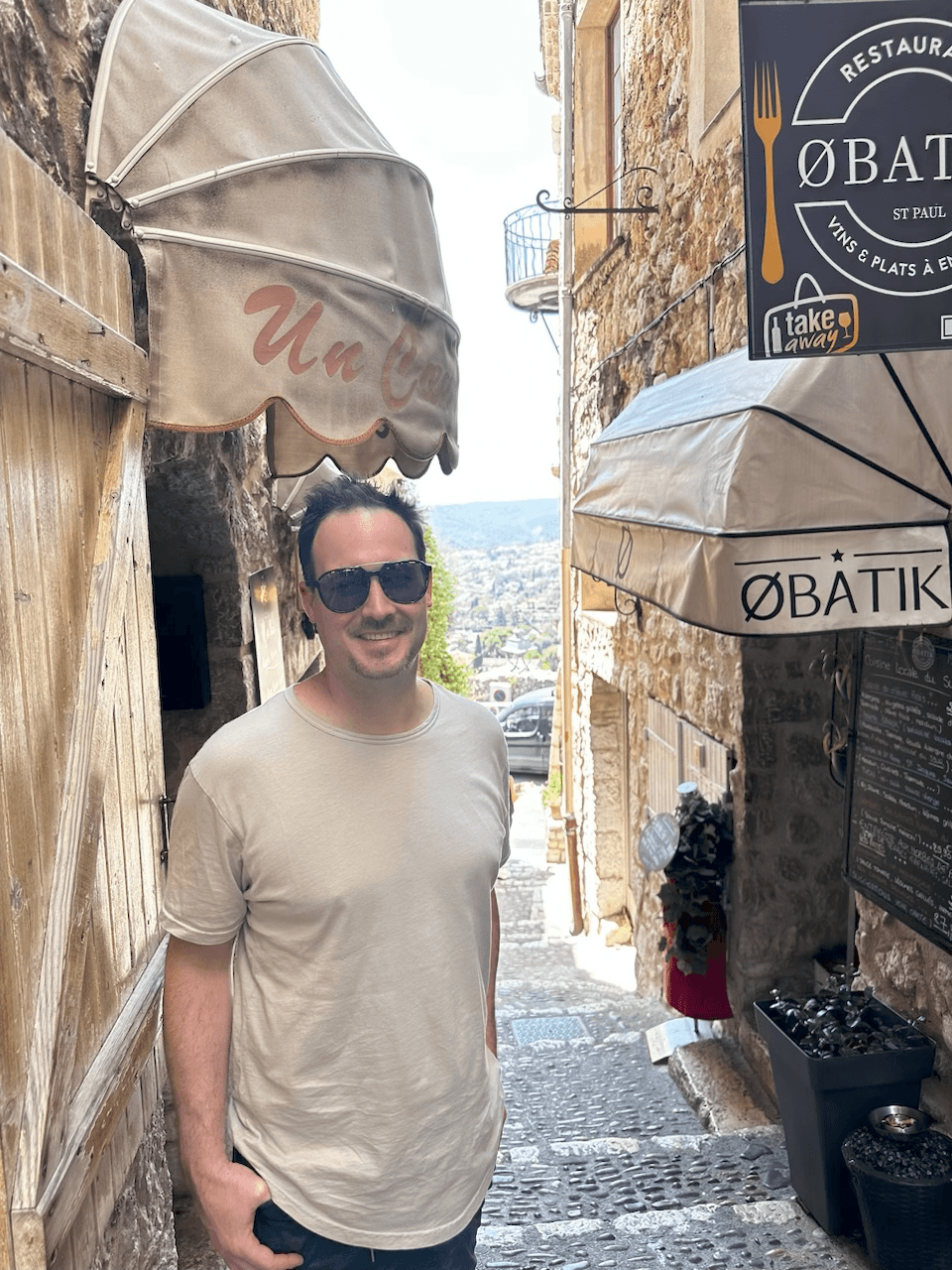 Person wearing sunglasses and a light-colored shirt standing on a narrow street with umbrellas and stone buildings.