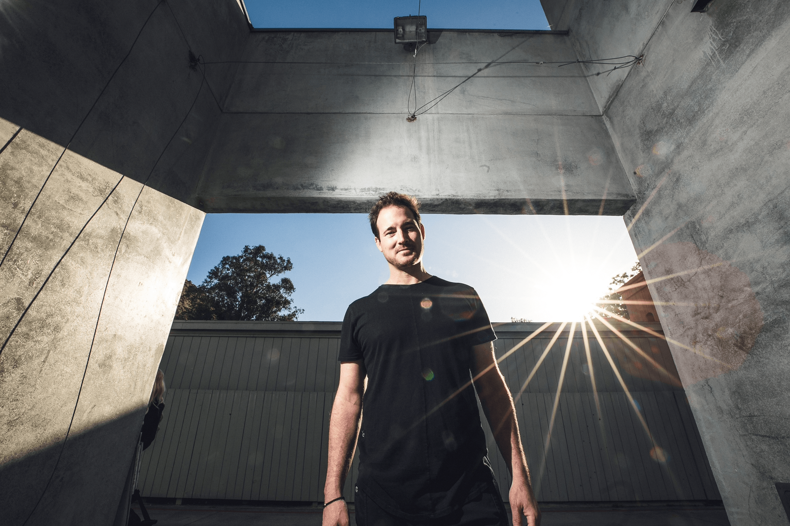 Young man standing in a concrete structure with sunlight shining behind him, blue sky visible above, smiling.