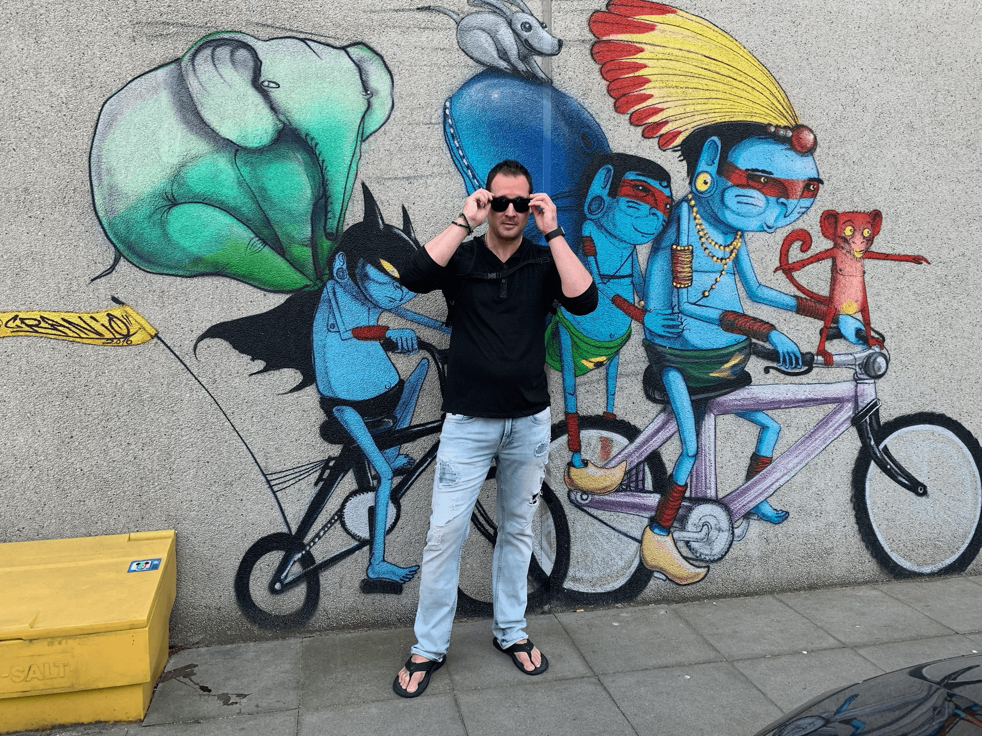 Person standing in front of colorful graffiti mural with characters riding a bicycle, holding sunglasses, wearing black shirt and light pants.