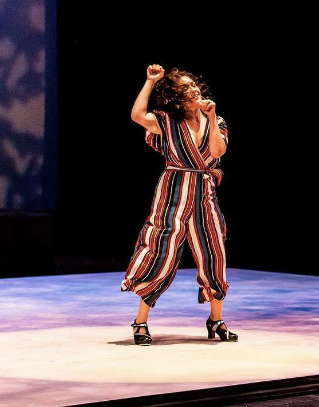 Person with curly hair dancing on stage, wearing striped jumpsuit and high heels, with one arm raised.
