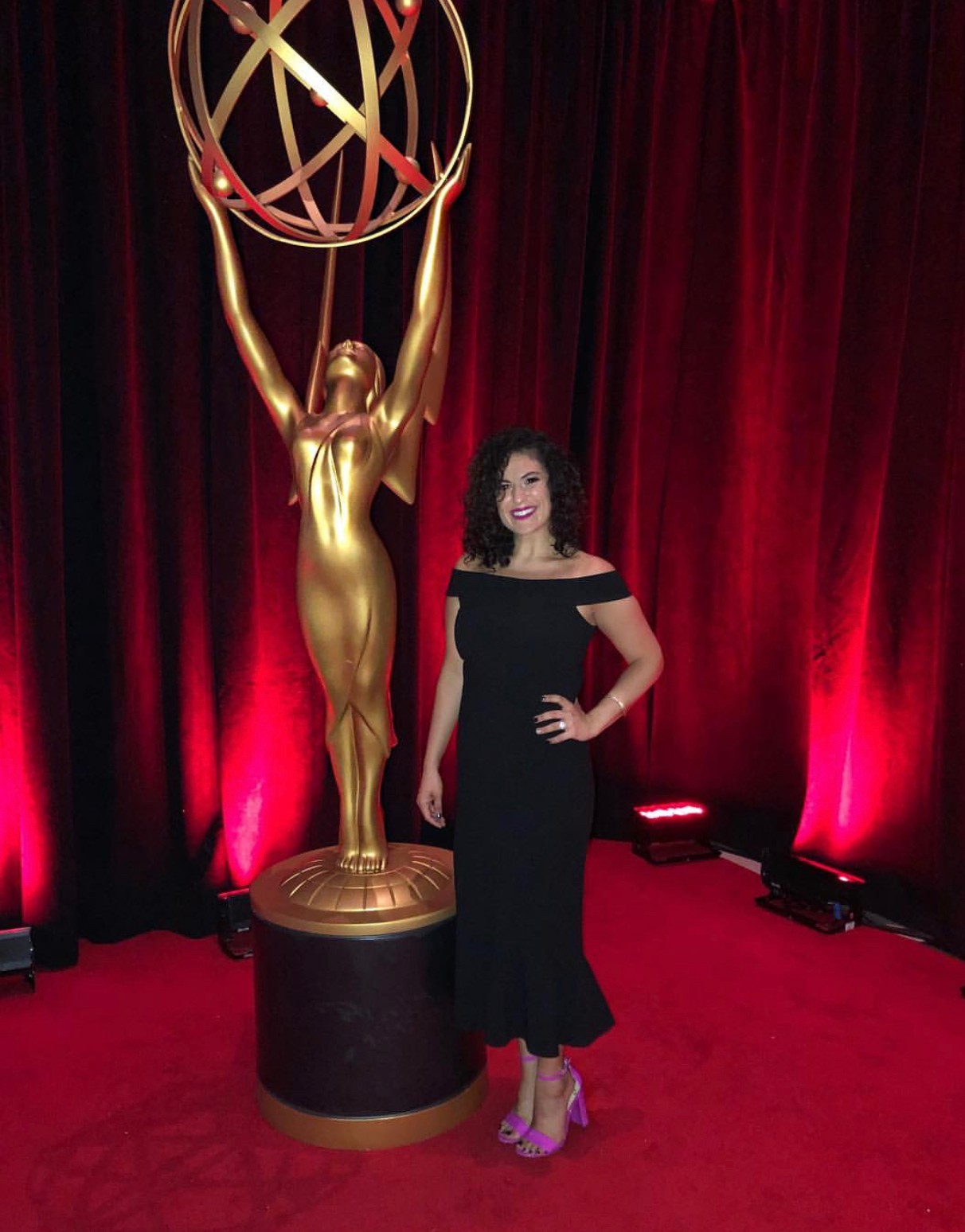 Woman in black dress standing next to a large gold Emmy statue on a stage with red curtains.