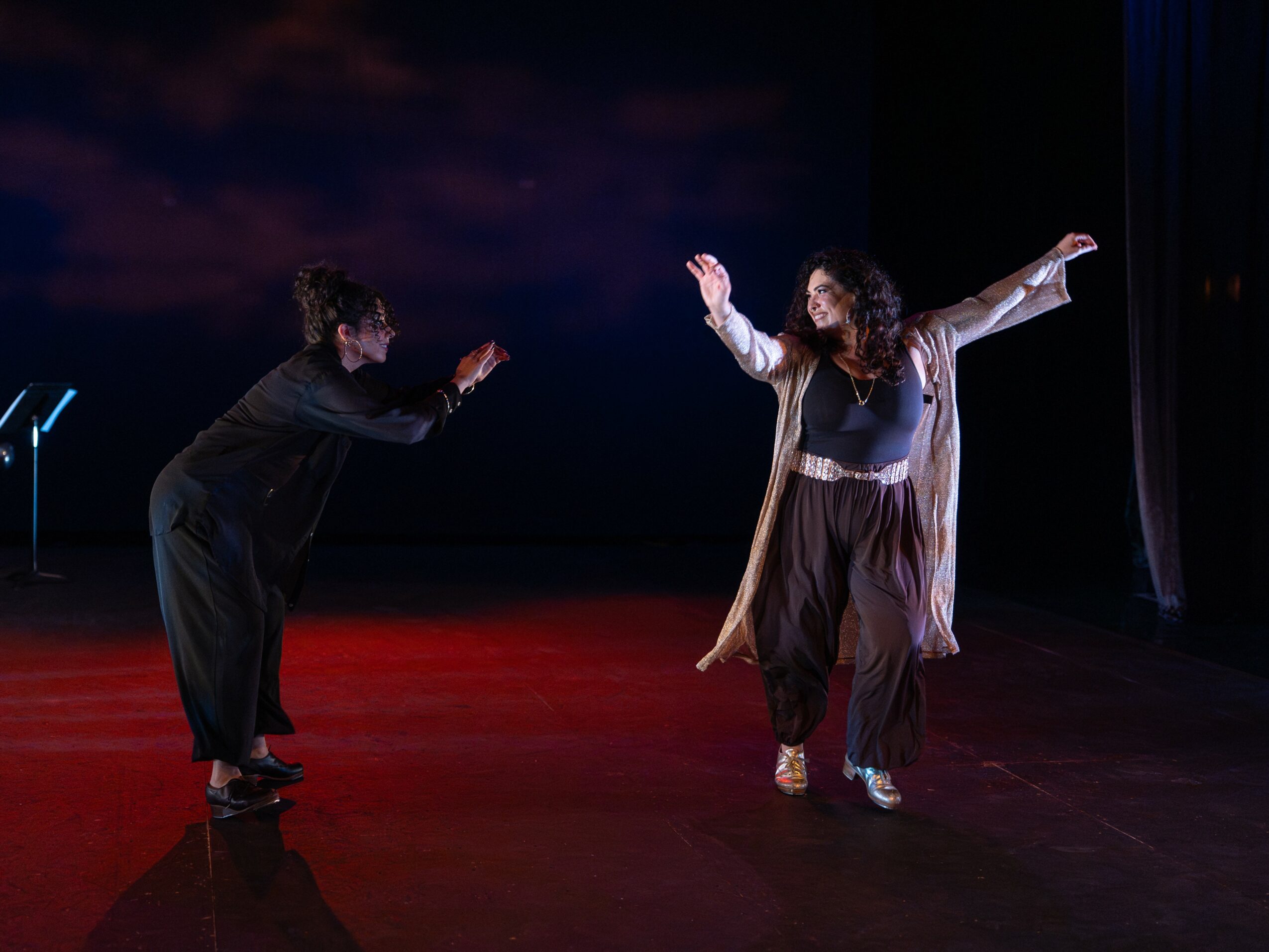 Two people dancing on stage, one with arms raised and the other bowing, with a dark background and red floor.