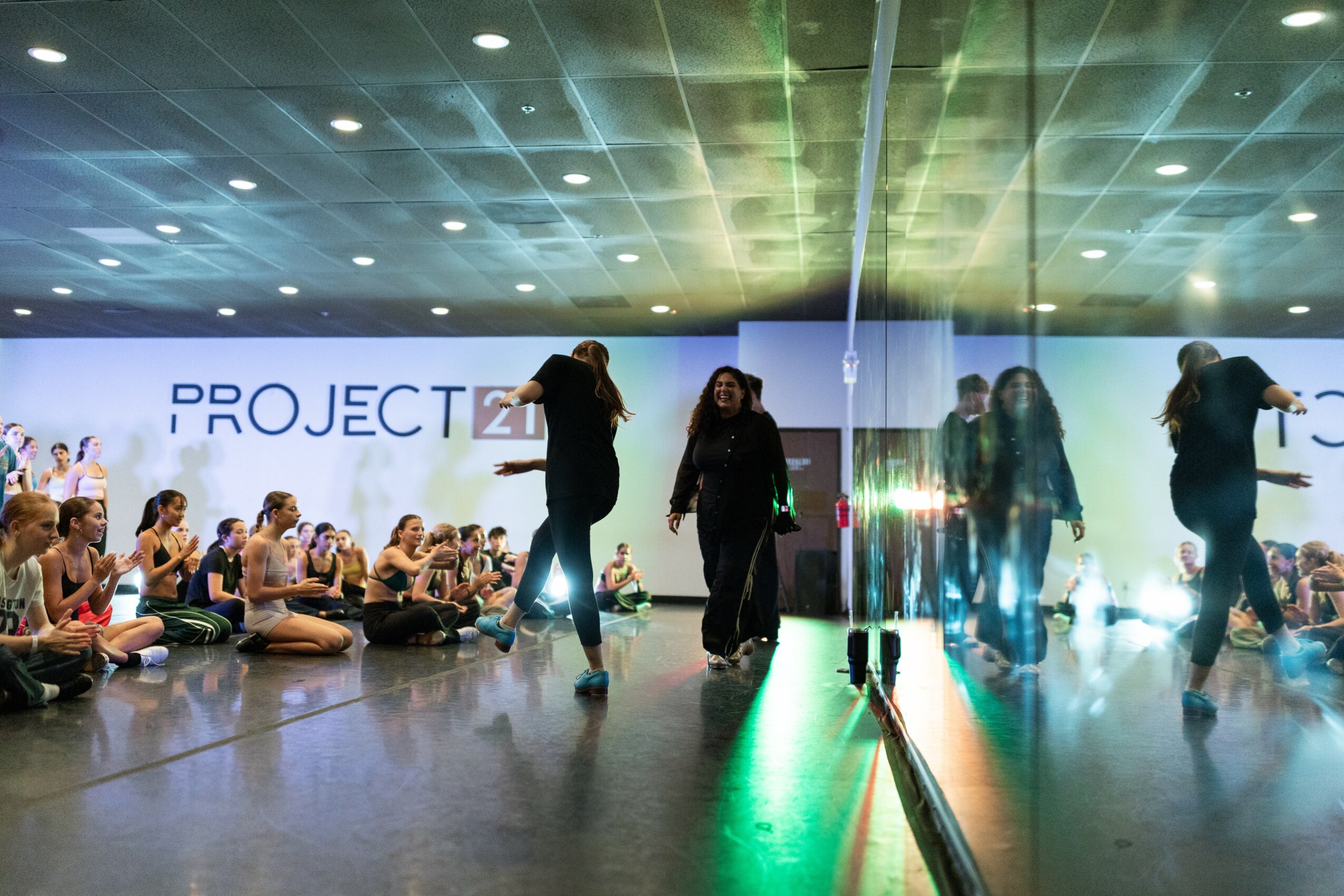 People walking in front of a large audience seated on the floor, with a sign reading 'PROJECT' in the background.