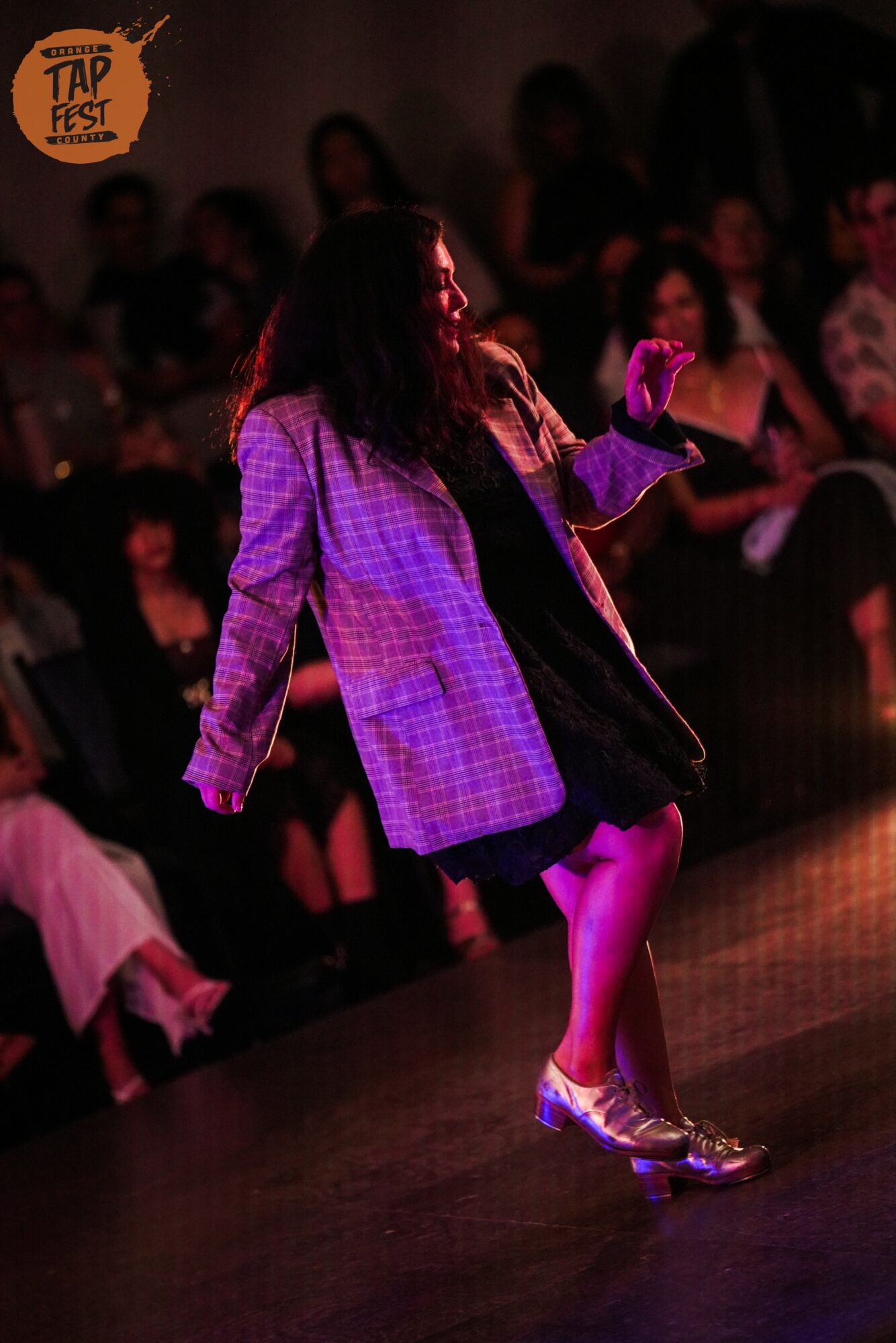Person dancing on stage with audience in background, wearing a checkered jacket and shiny shoes, illuminated by stage lighting.