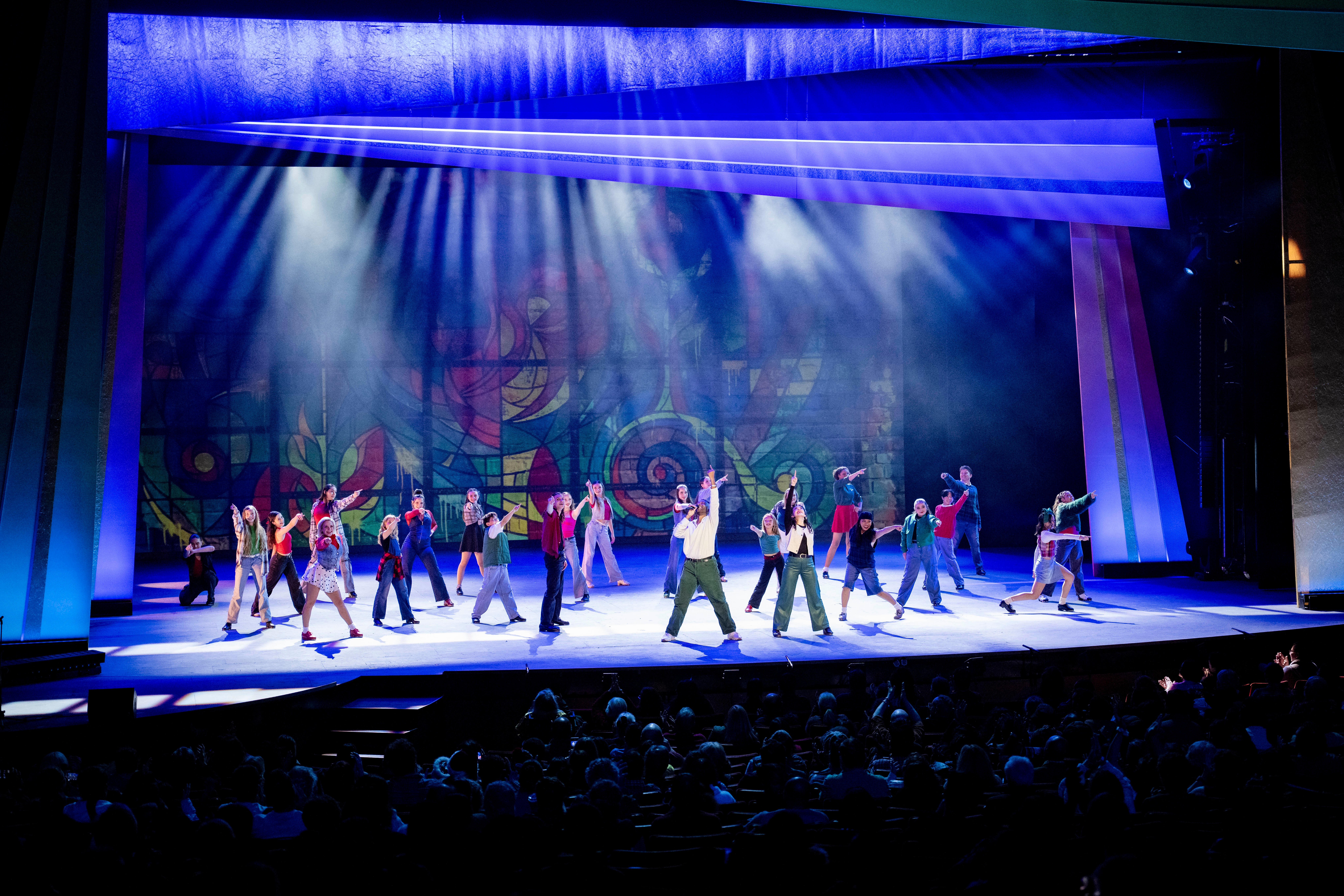 Group of performers on stage with colorful backdrop, stage lights shining down, audience visible in foreground.