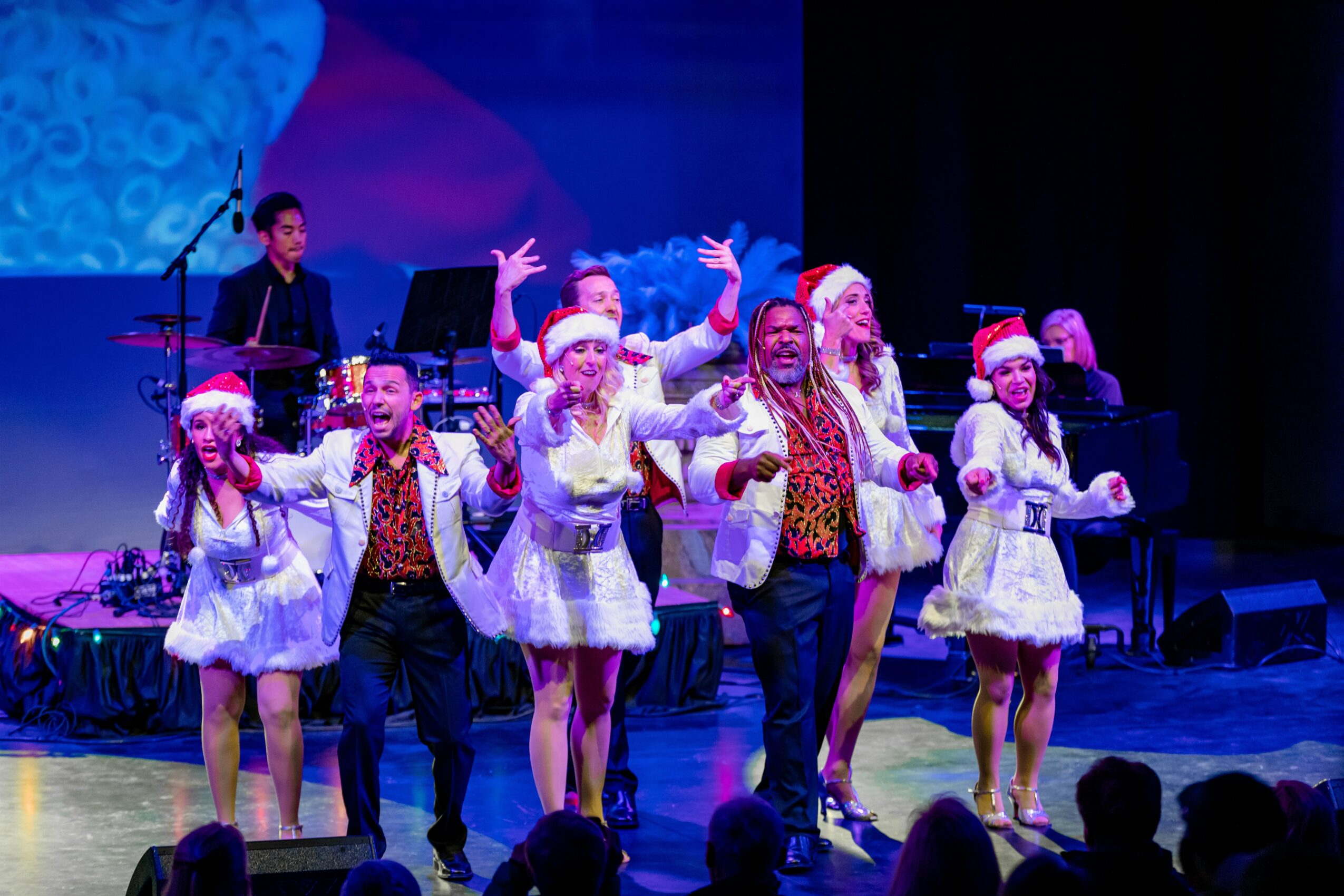 Group of performers on stage wearing festive costumes, some with Santa hats, dancing and singing with musicians in the background.