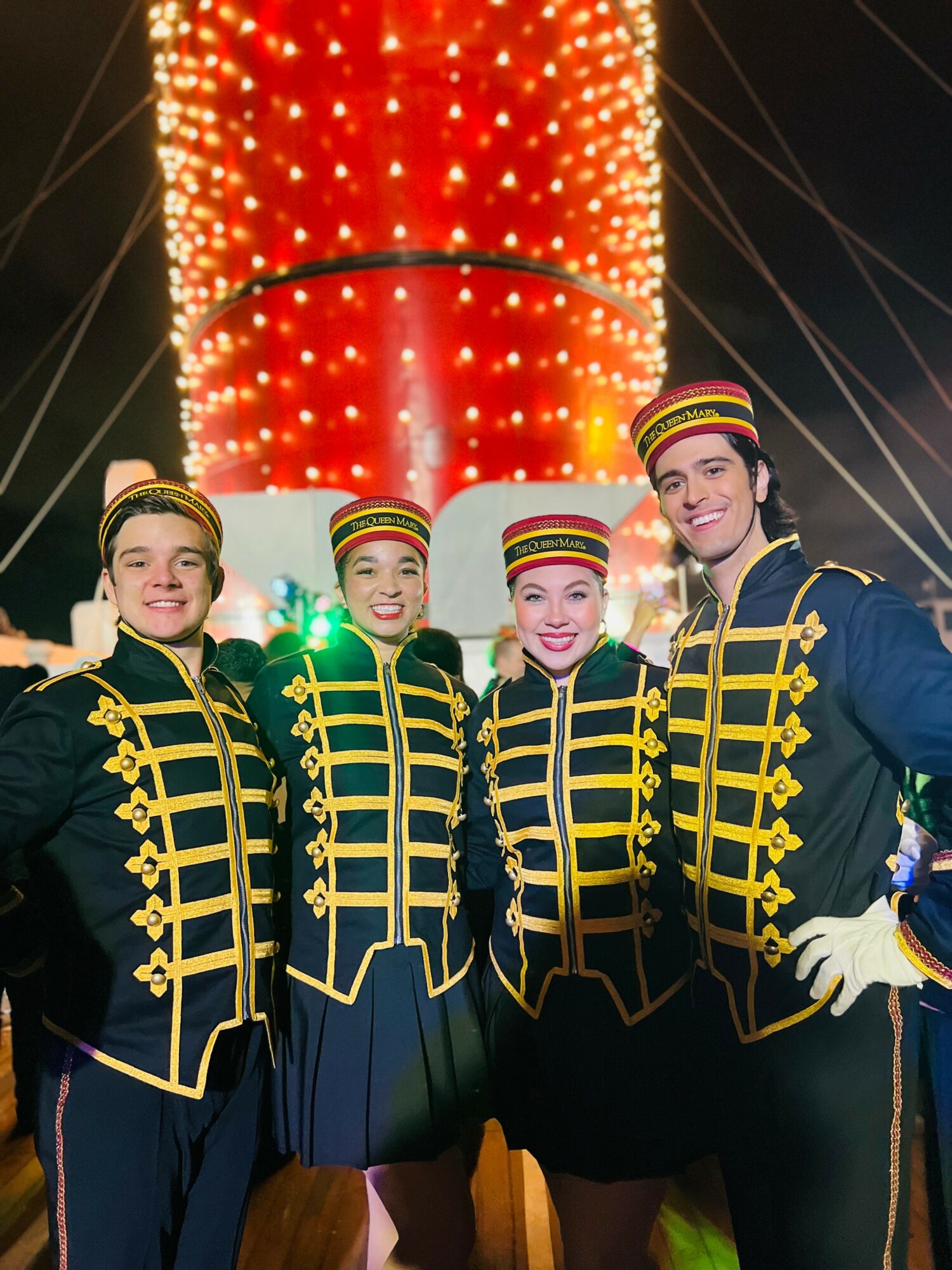 Four performers in matching costumes stand in front of a brightly lit carnival ride at night.