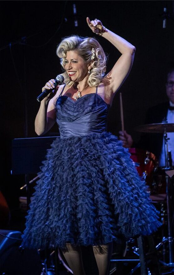 Woman singing into a microphone, wearing a dark blue dress with a ruffled skirt, smiling, with a band in the background.