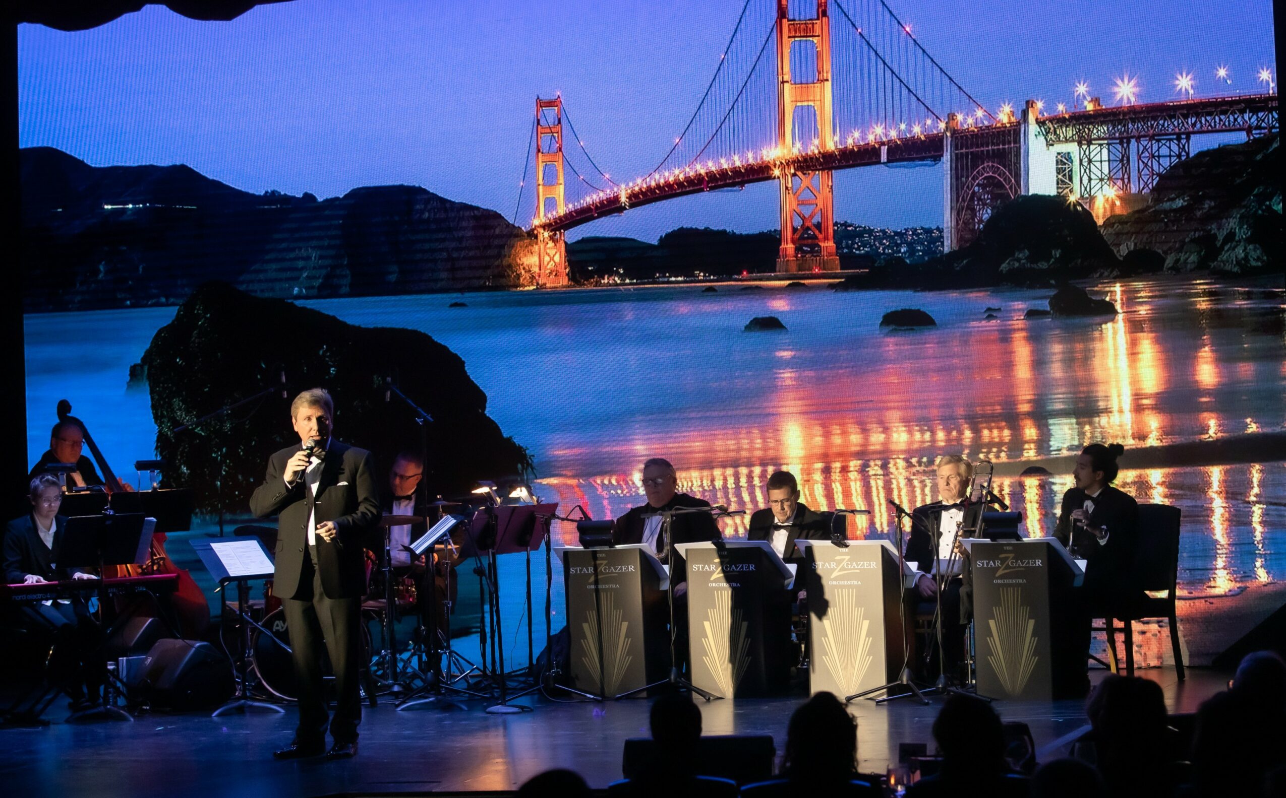 Musicians perform on stage with a cityscape featuring a bridge illuminated at night in the background.