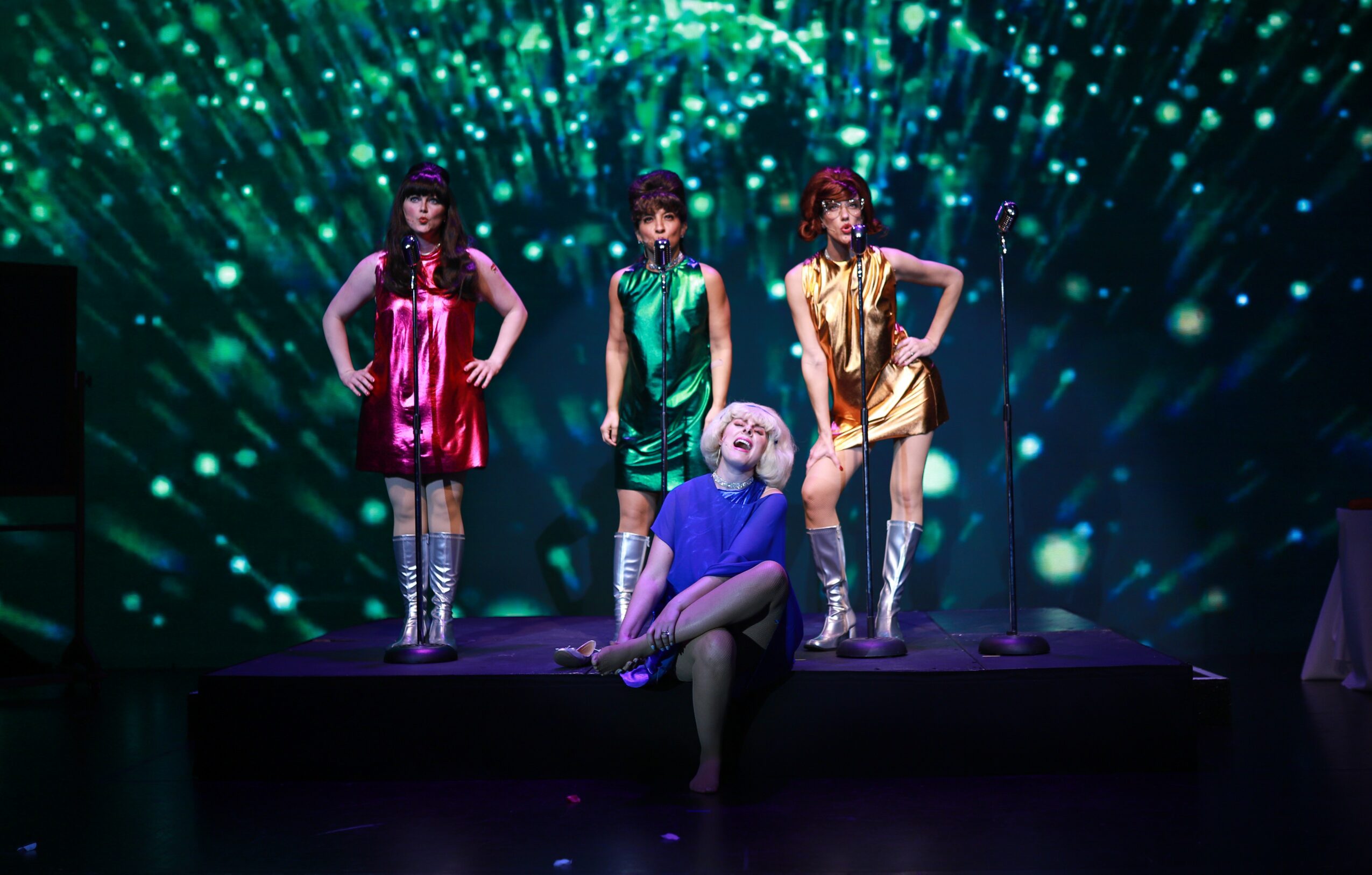 Four performers on stage with colorful costumes and a person sitting in front, illuminated by green and blue lights.