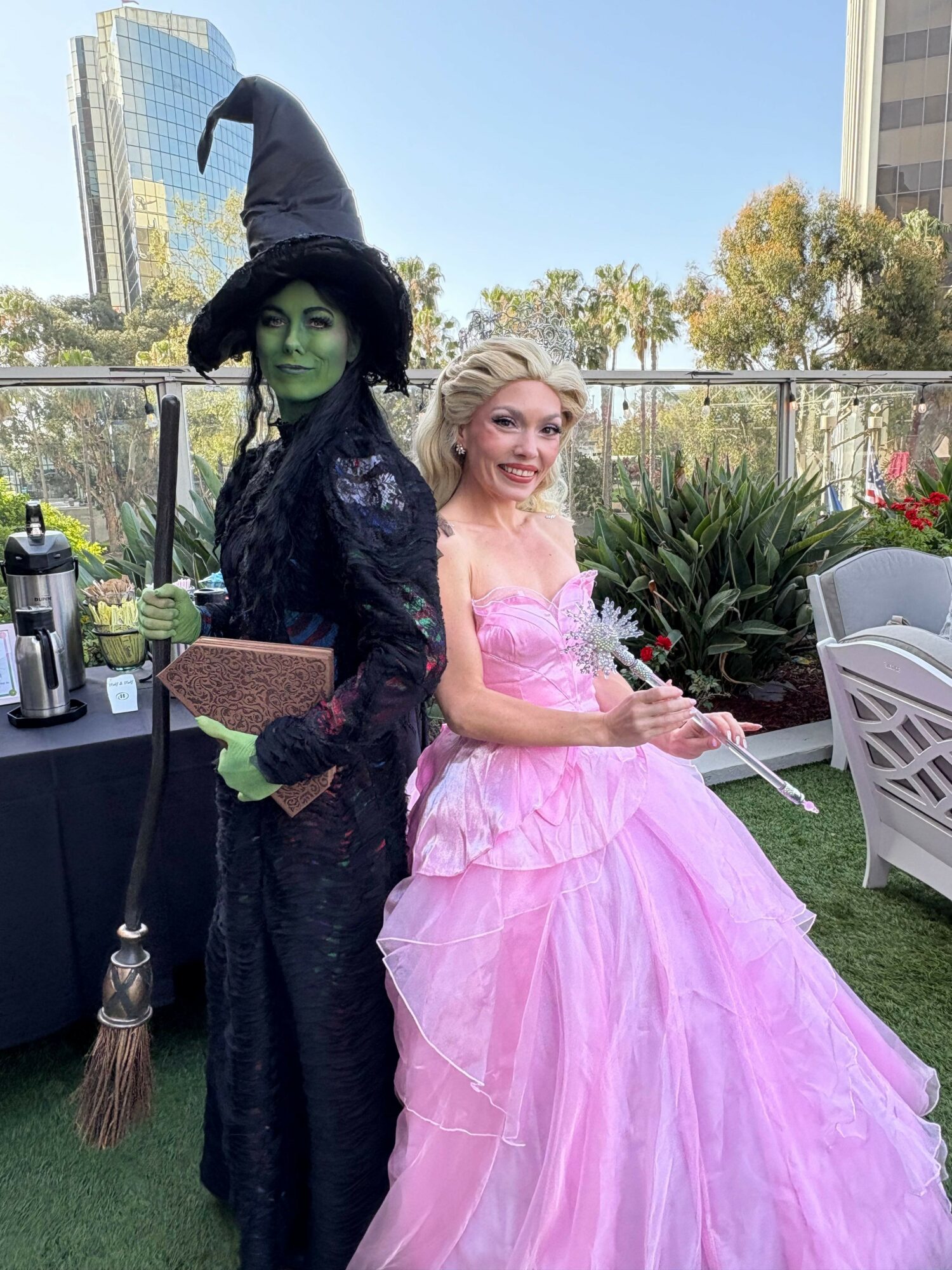 Person dressed as a witch with green face paint and a black hat, standing next to a woman in a pink gown outdoors.