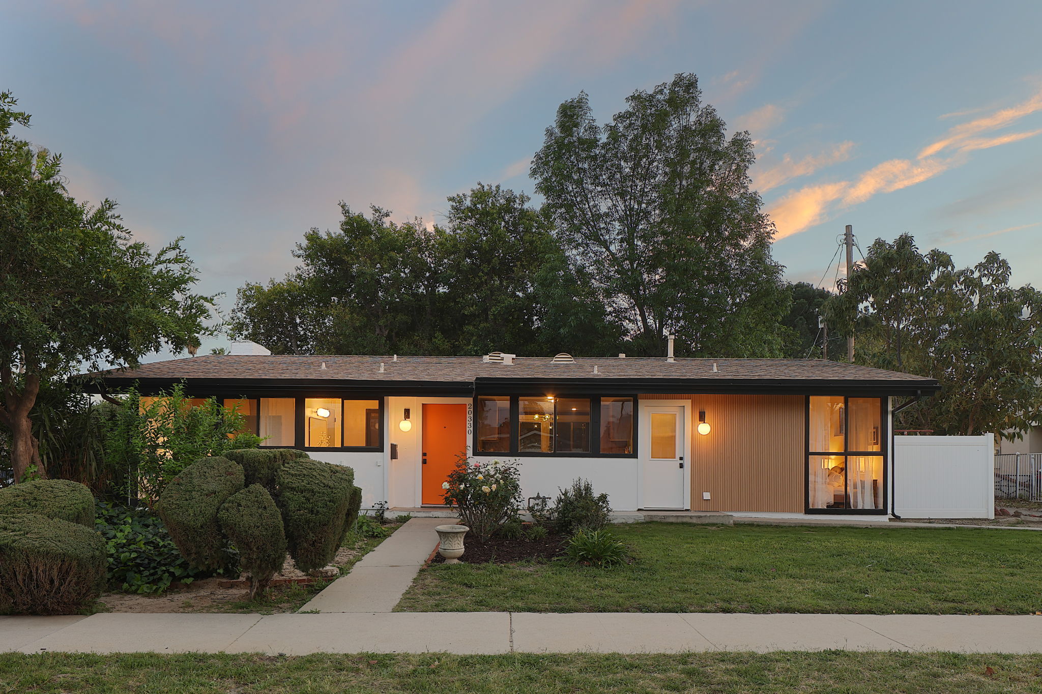 Single-story house with a flat roof, large windows, and a front door, surrounded by trees and a lawn.
