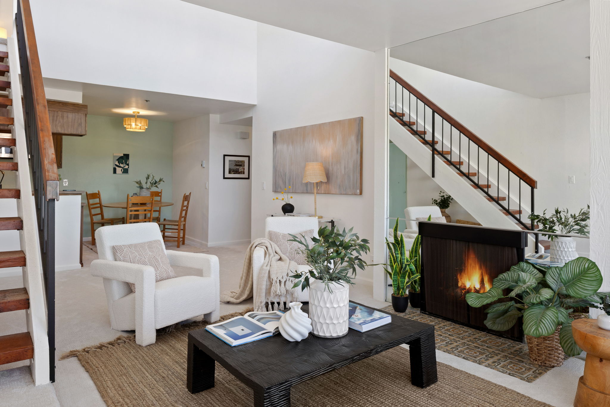 Living room with white armchair, black coffee table, fireplace, staircase, and potted plants, with dining area in background.