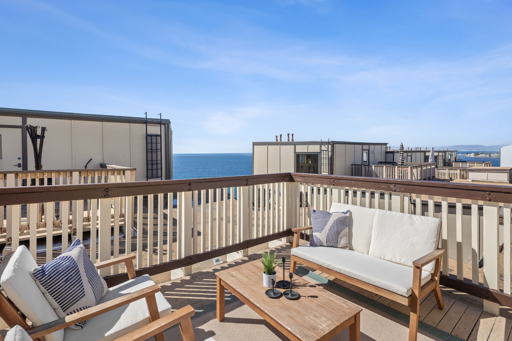 Rooftop terrace with outdoor furniture, wooden railing, and ocean view under a blue sky.