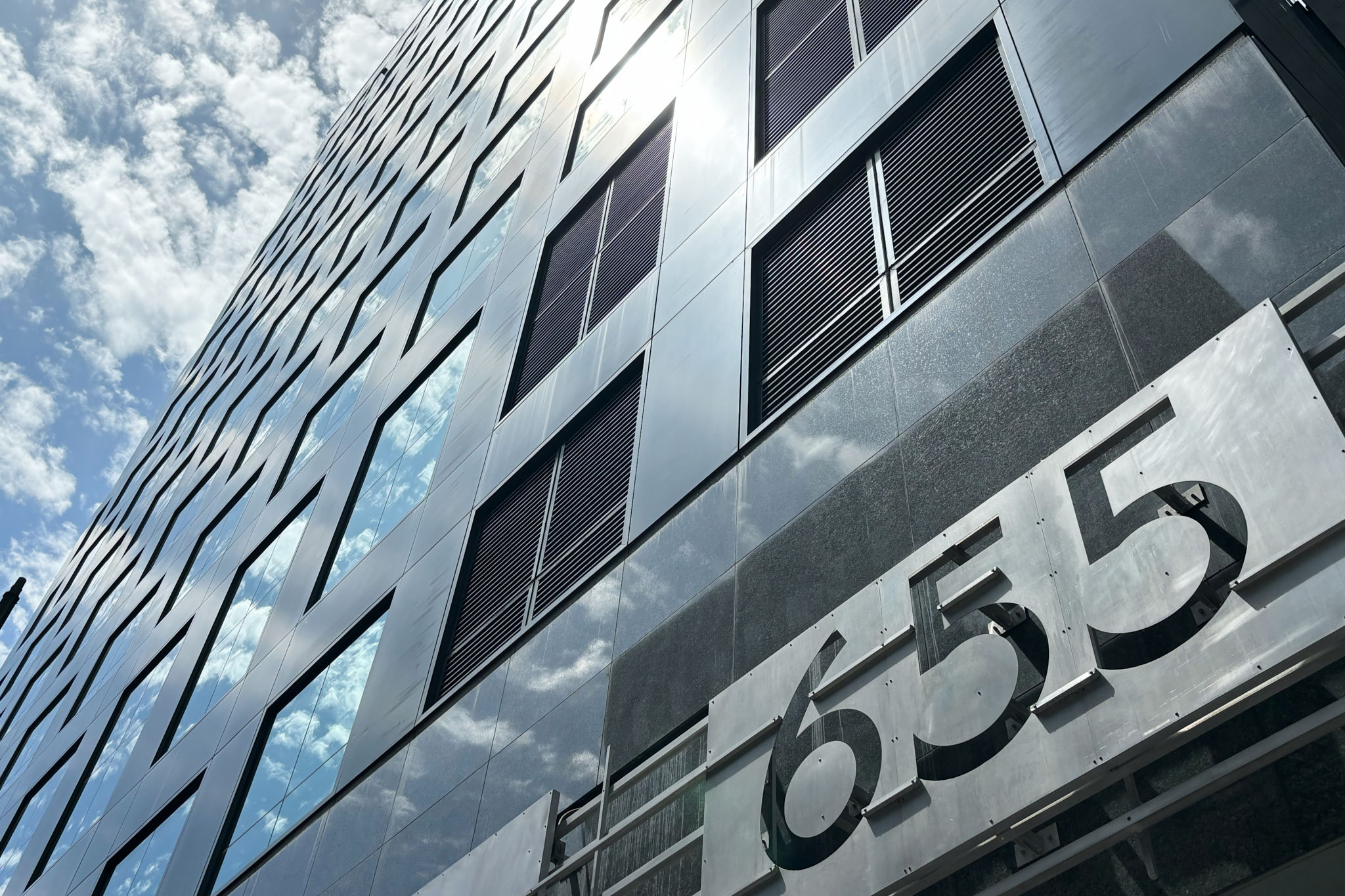 Modern building with reflective glass windows and a street number 655 on the facade, sky with clouds reflected in windows.