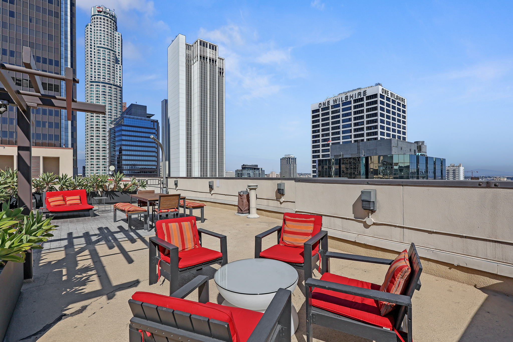 Rooftop terrace with red chairs and a round table, city skyline with tall buildings in the background, blue sky.