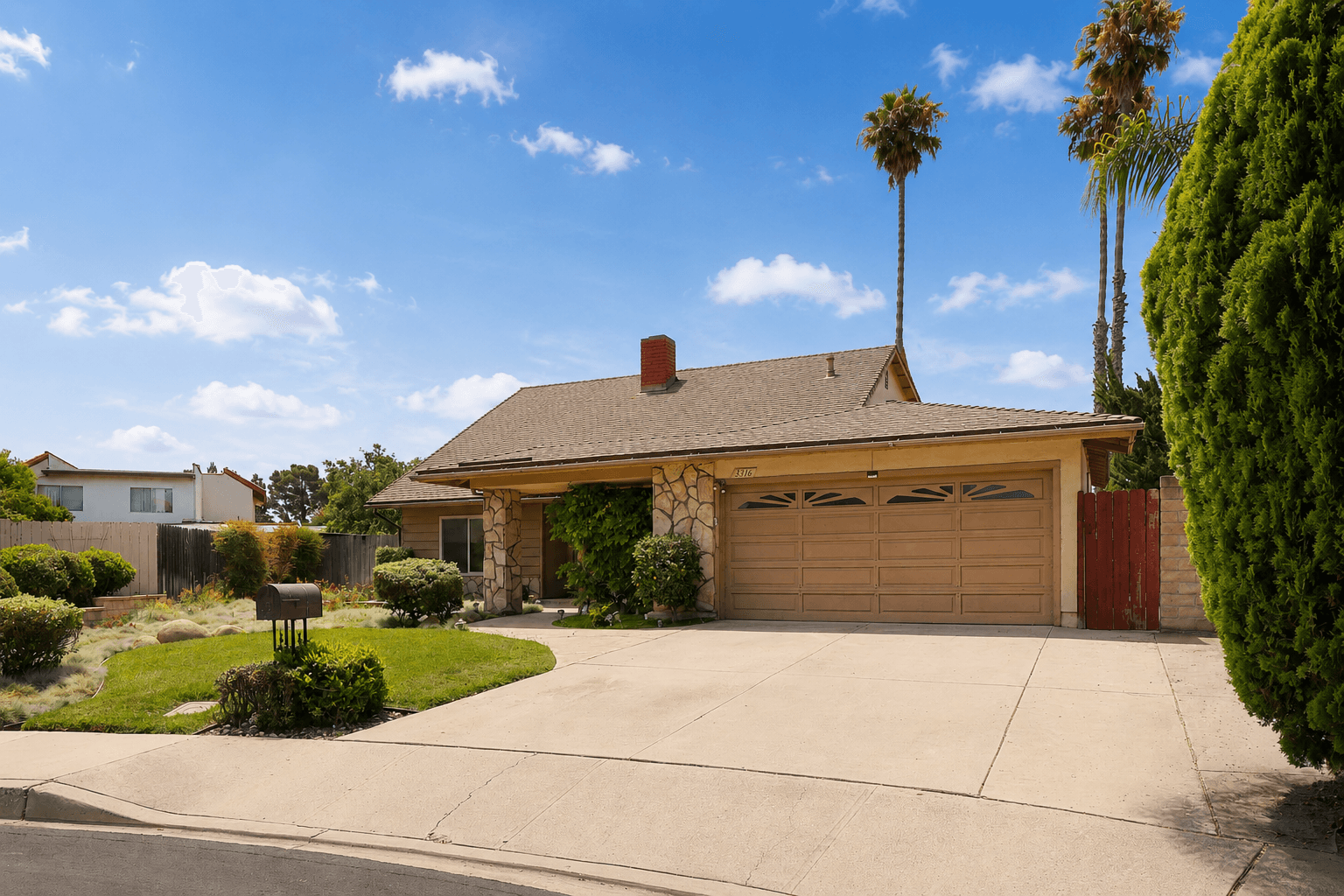 Single-story house with a brown garage door, surrounded by a well-maintained lawn and tall palm trees, under a blue sky.