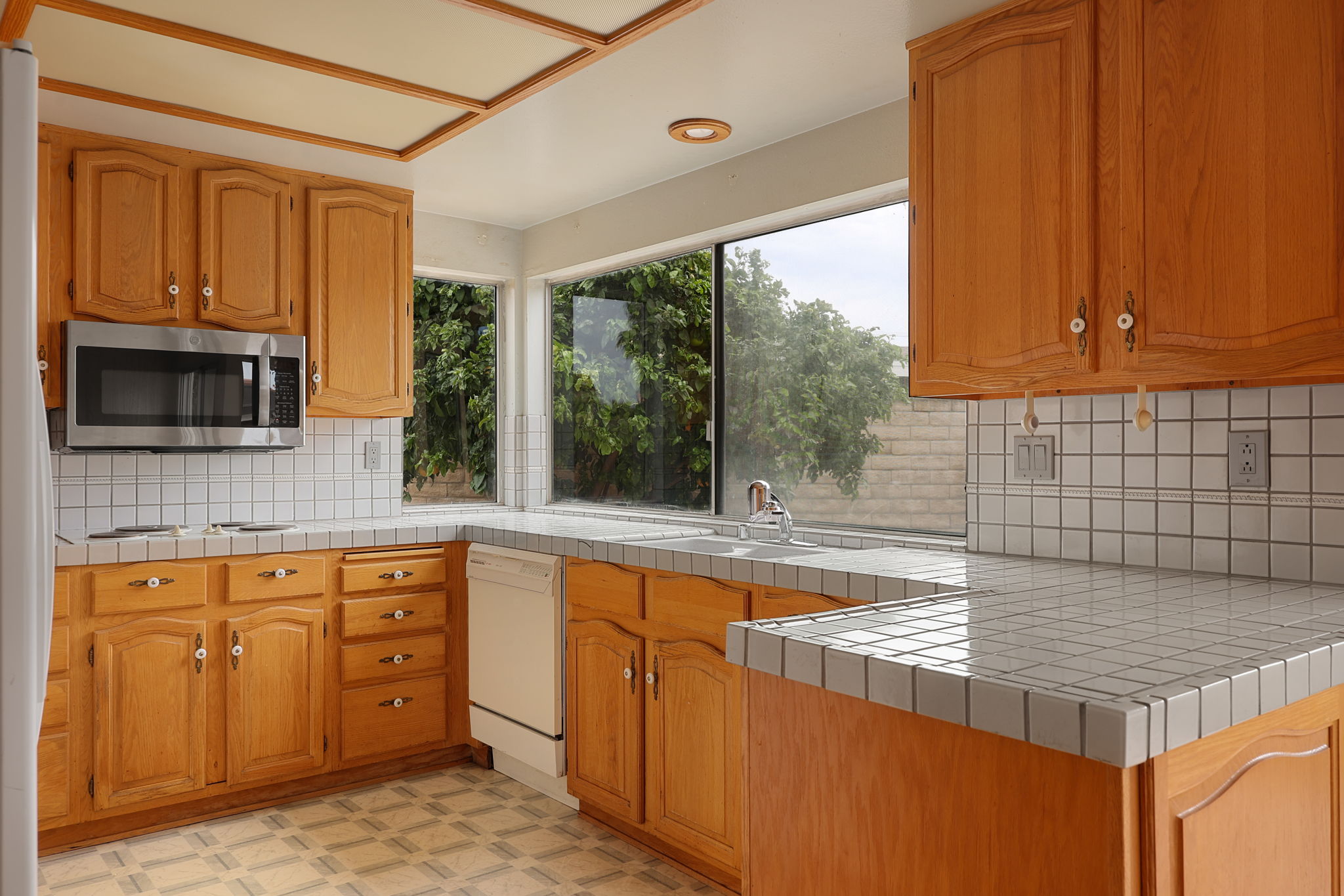 Kitchen with wooden cabinets, white tiled countertops, and a large window overlooking trees, with a microwave and sink visible.