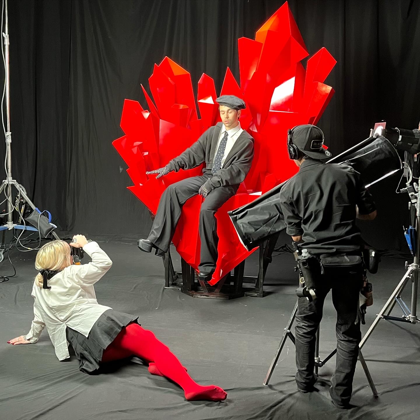 Person in graduation gown and cap seated on a red abstract throne, with a child taking photos and a camera operator nearby.