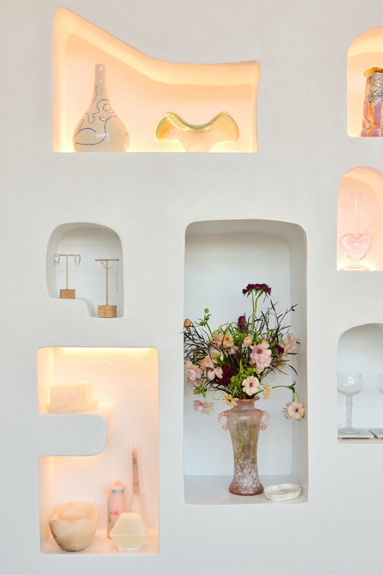 Vase with pink and white flowers on a white shelf, surrounded by decorative objects and illuminated niches.